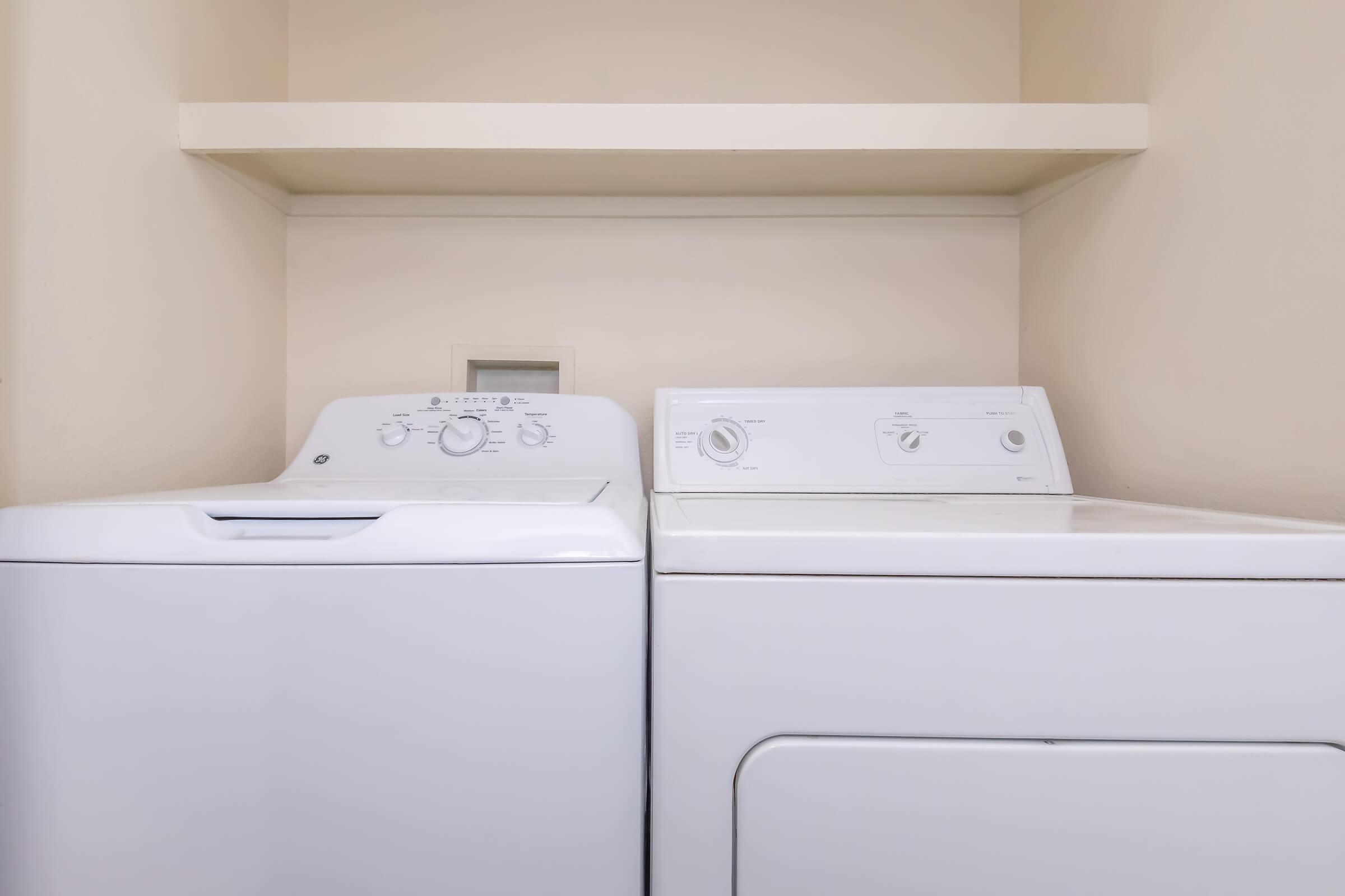 A compact laundry area featuring a white washing machine and a white dryer side by side, with a beige wall in the background and a shelf above them for storage. The setting appears clean and organized, suitable for a home or apartment laundry space.