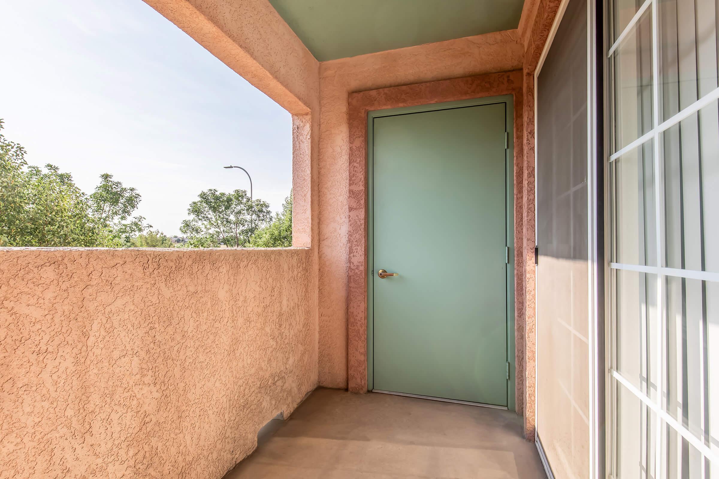 A balcony with a peach-colored textured wall and a green door. The space is open with a view of trees outside. There's natural light brightening the area, and a railing partially visible on one side. The overall aesthetic is clean and simple, offering a tranquil outdoor space.