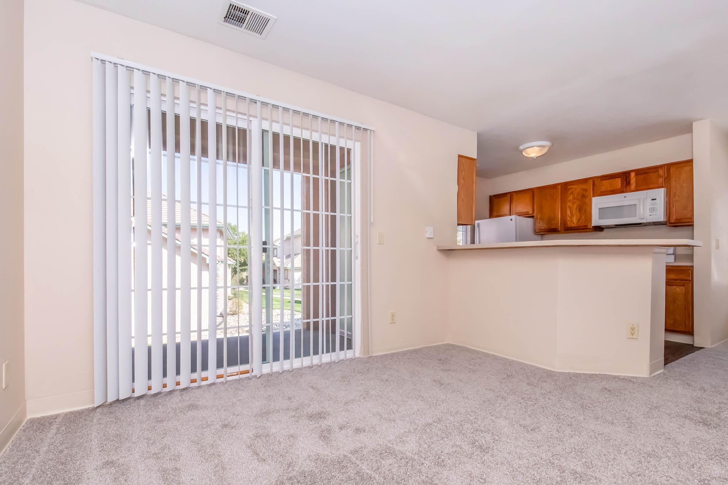 A spacious living area with light carpet and large sliding glass doors leading to a patio. The room has beige walls and a small breakfast bar that connects to a kitchen with wooden cabinets. Natural light fills the space, creating an inviting atmosphere.