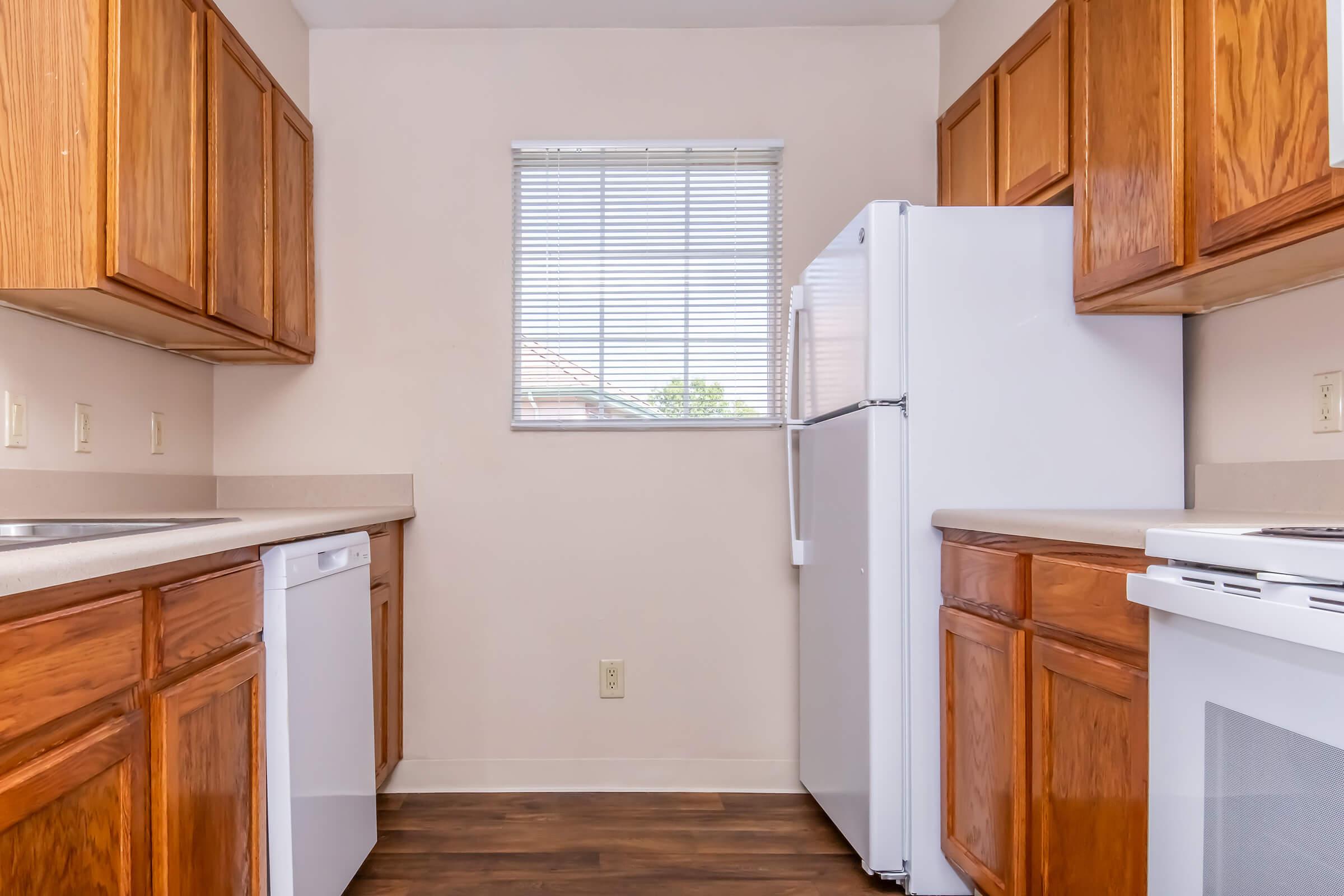 A bright, clean kitchen featuring wooden cabinets, a white refrigerator, and a white dishwasher. The room has a window with blinds allowing natural light to enter, and there is a light-colored countertop. The flooring is dark wood, creating a warm contrast with the cabinetry.