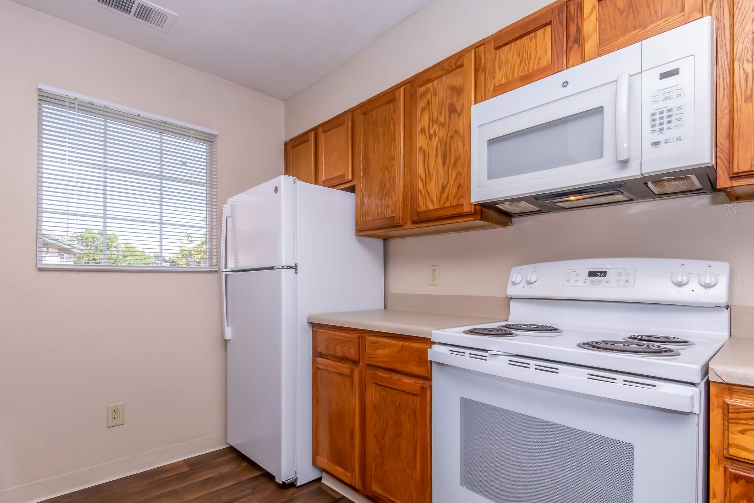 A compact kitchen featuring wooden cabinets, a white refrigerator, and an oven with a stovetop. The kitchen has a window with blinds that lets in natural light. The countertops are beige, providing a neutral backdrop to the appliances and cabinetry.