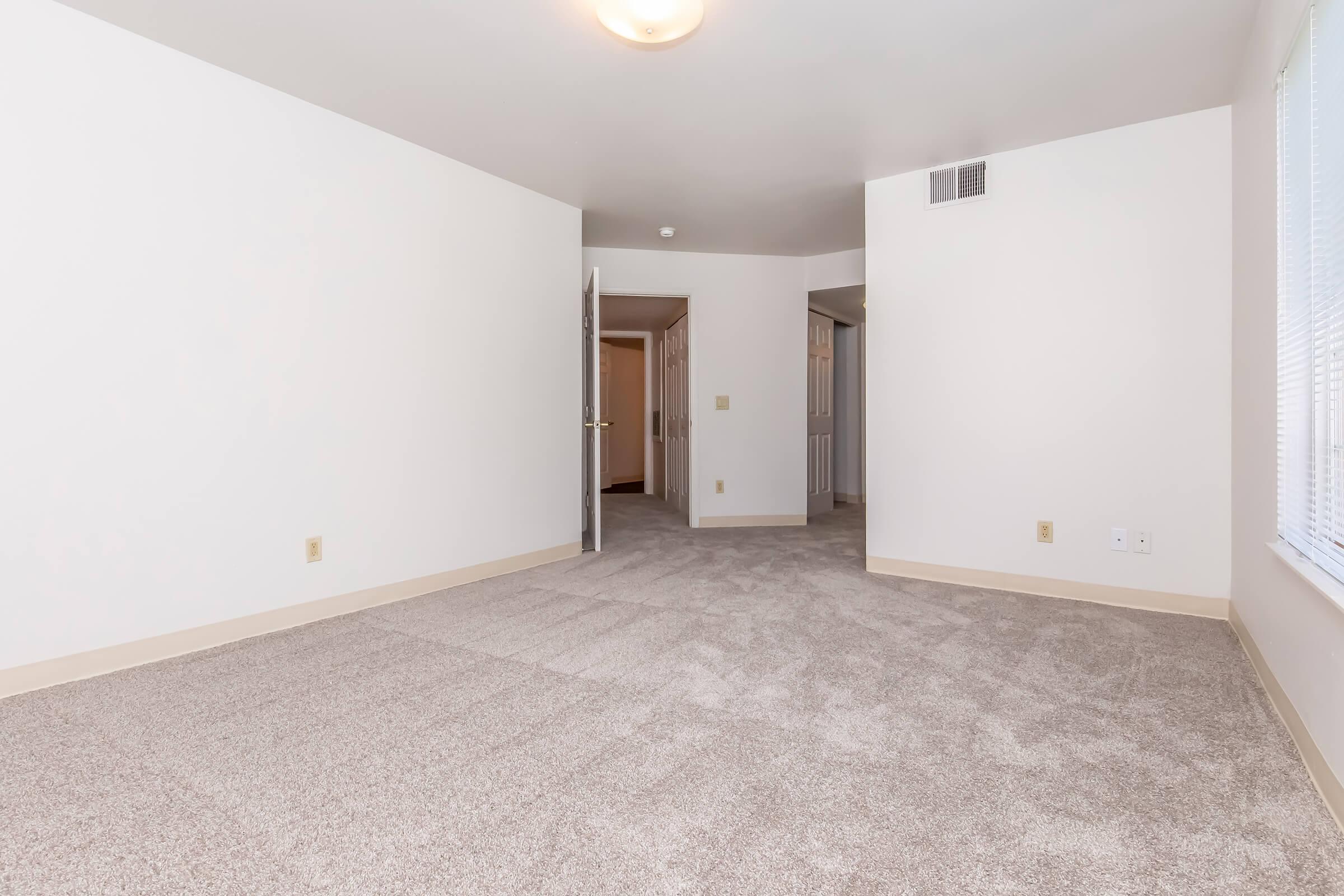 A spacious, unfurnished room with light gray carpet, neutral walls, and a ceiling light fixture. The room features a doorway leading to another room, and a large window allowing natural light to enter. The atmosphere is bright and open, suitable for various design options.
