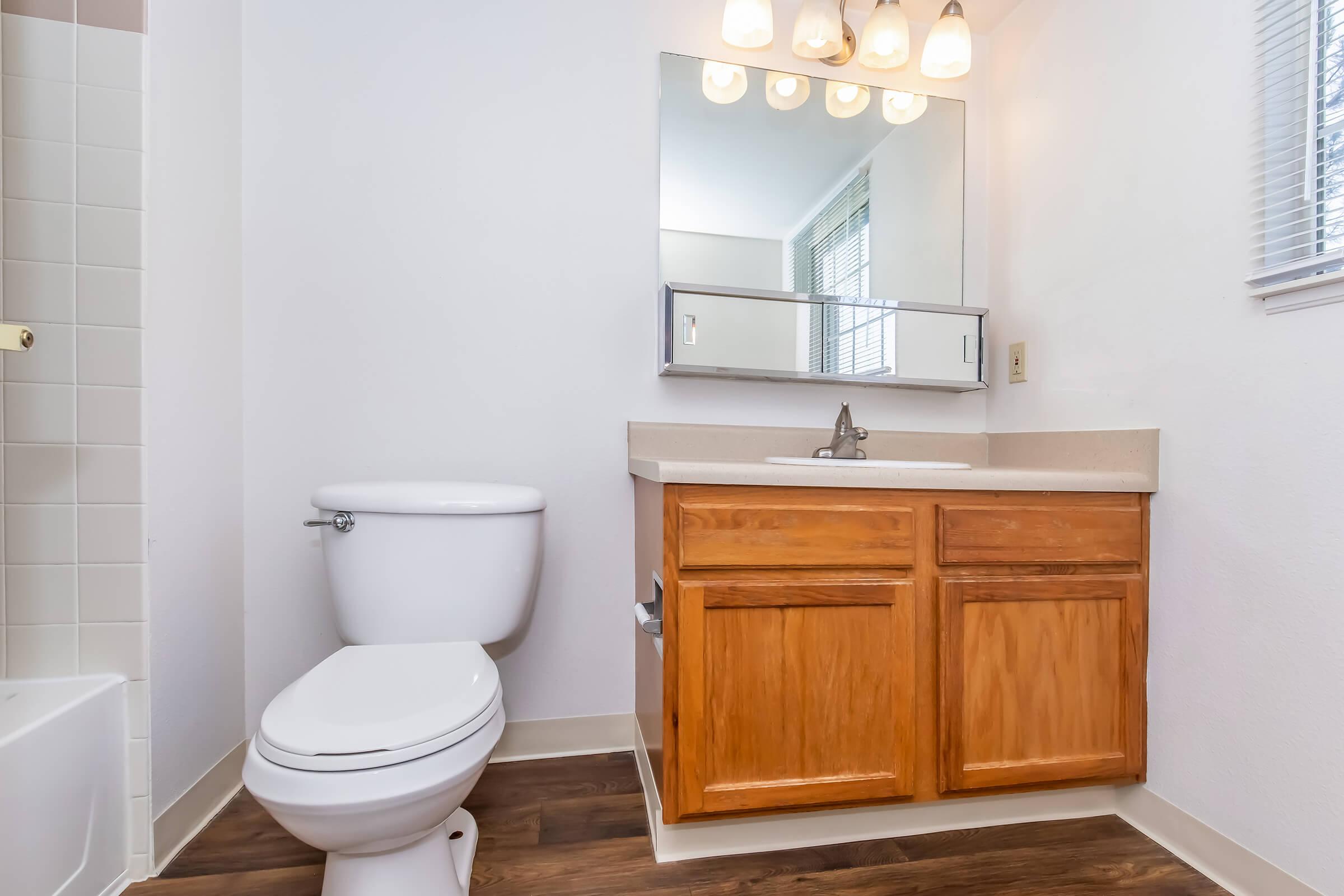 A clean, modern bathroom featuring a white toilet, a wooden vanity with a sink, and a mirror with light fixtures above. The room has neutral walls and flooring, with a window allowing natural light. A bathtub is partially visible on the left side.