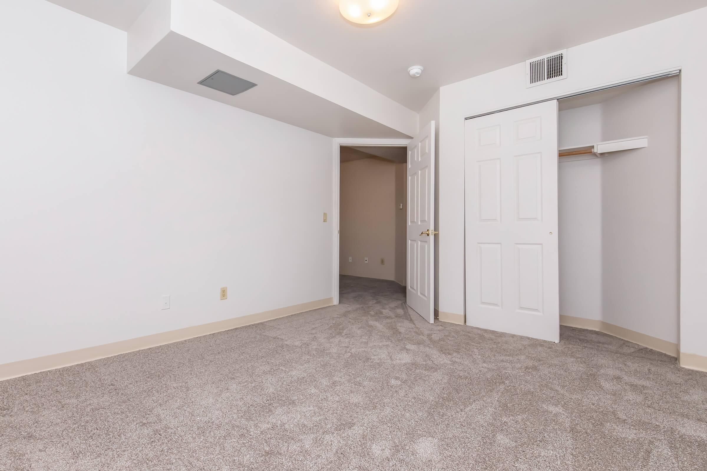 A spacious, empty room with light beige carpet and white walls. A closed closet with double doors is visible, along with a doorway leading to another room. The ceiling features a light fixture, and the overall ambiance is bright and inviting.
