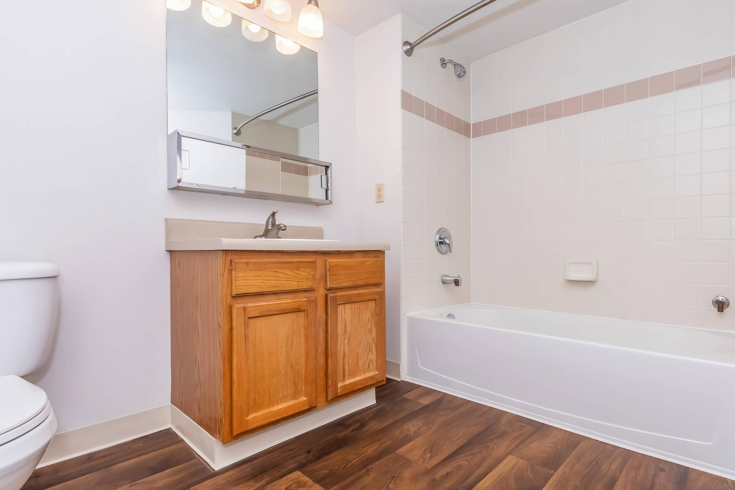 A clean, modern bathroom featuring a white bathtub with a showerhead, a wooden vanity with two cabinets, a mirror above the sink, and a toilet. The floor has a dark wooden appearance, while the walls have light-colored tiles with a pink accent stripe. Bright lighting from overhead fixtures illuminates the space.
