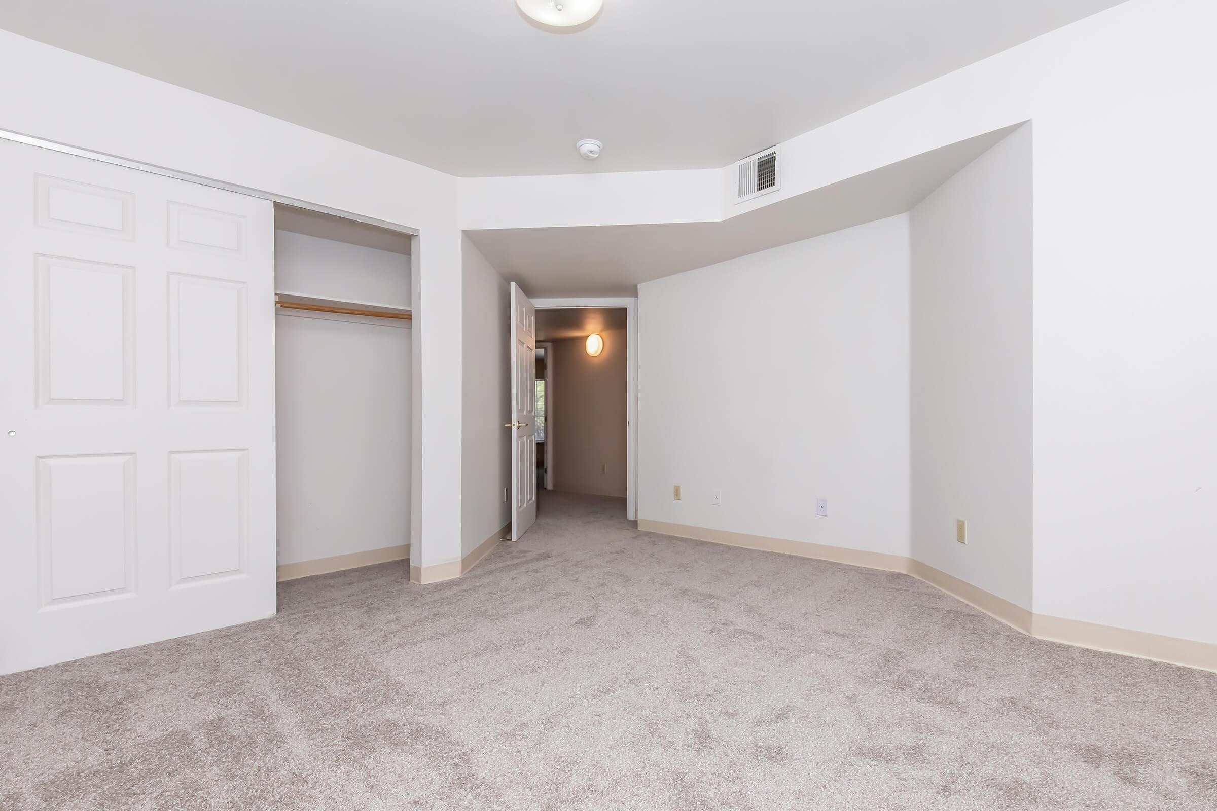 A spacious, empty room with beige carpet flooring and light-colored walls. There is a closet with sliding doors on the left side, and a doorway leading to another area on the right. A ceiling light fixture provides illumination, creating a bright and open atmosphere.
