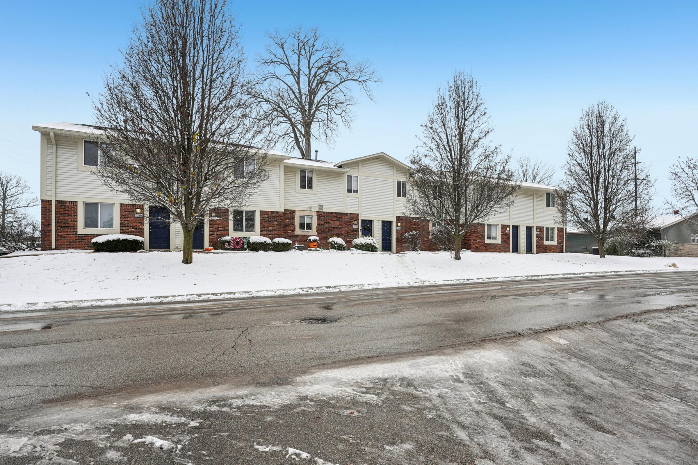 A snowy residential neighborhood featuring several two-story apartment buildings with brick and siding exteriors. Bare trees line the street, and a slight snow cover can be seen on the ground. The sky is clear with a blue hue, indicating a cold winter day.