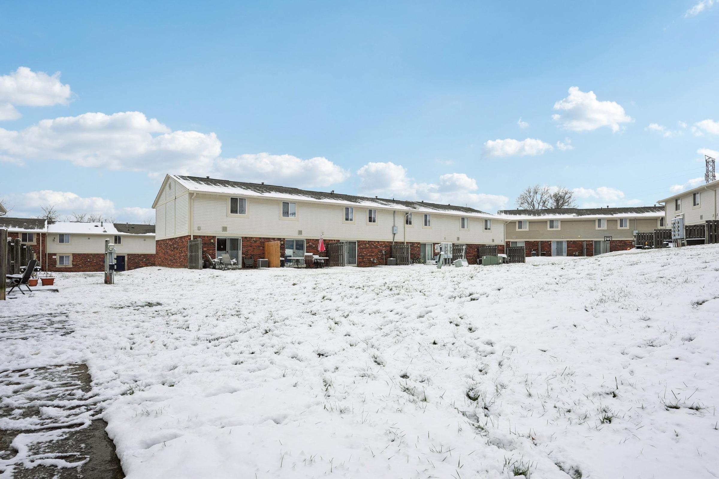 A winter scene featuring a row of residential buildings, mostly two-story townhouses with white exteriors and brick accents. The ground is covered in a layer of freshly fallen snow, and the sky is a clear blue with scattered clouds. Some outdoor furniture and recreational equipment are partially visible.