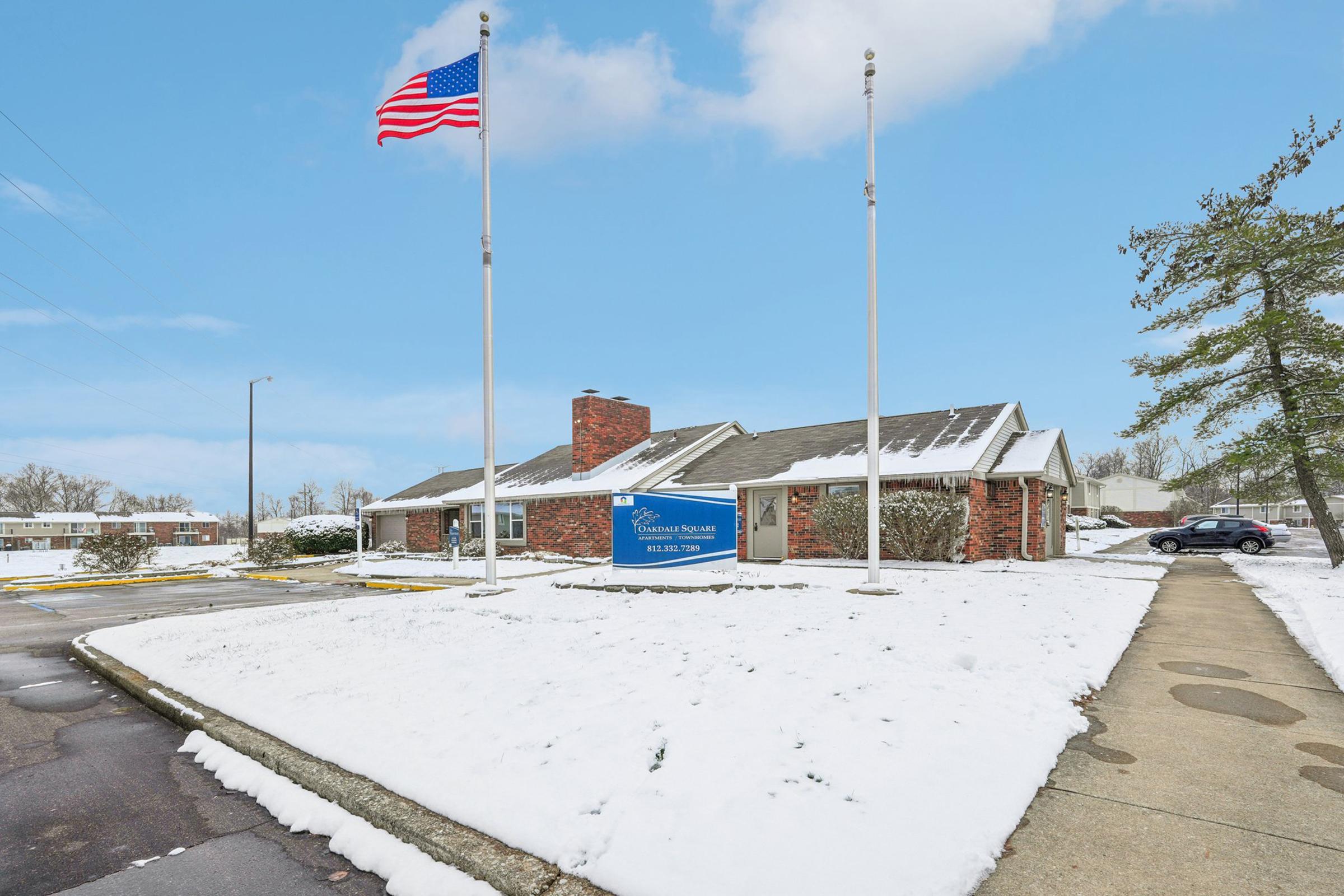 A one-story brick building with a sloped roof, surrounded by a layer of snow. Two flagpoles display an American flag and a state flag. There are trees in the background and a snowy sidewalk leading to the entrance. The sky is clear with some clouds. A sign in front indicates the building's name.