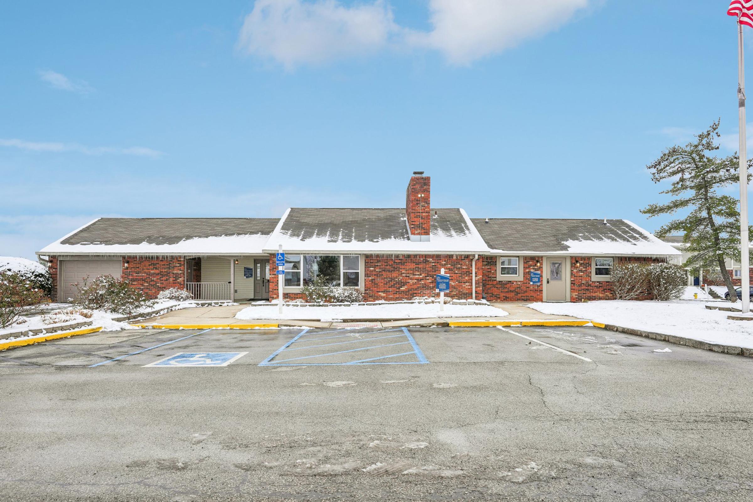 A single-story brick building with a snowy roof and drive-up parking lot. The front features a chimney and two sets of entryways flanked by blue parking signs. A clear blue sky with scattered clouds is in the background, enhancing the winter scene.