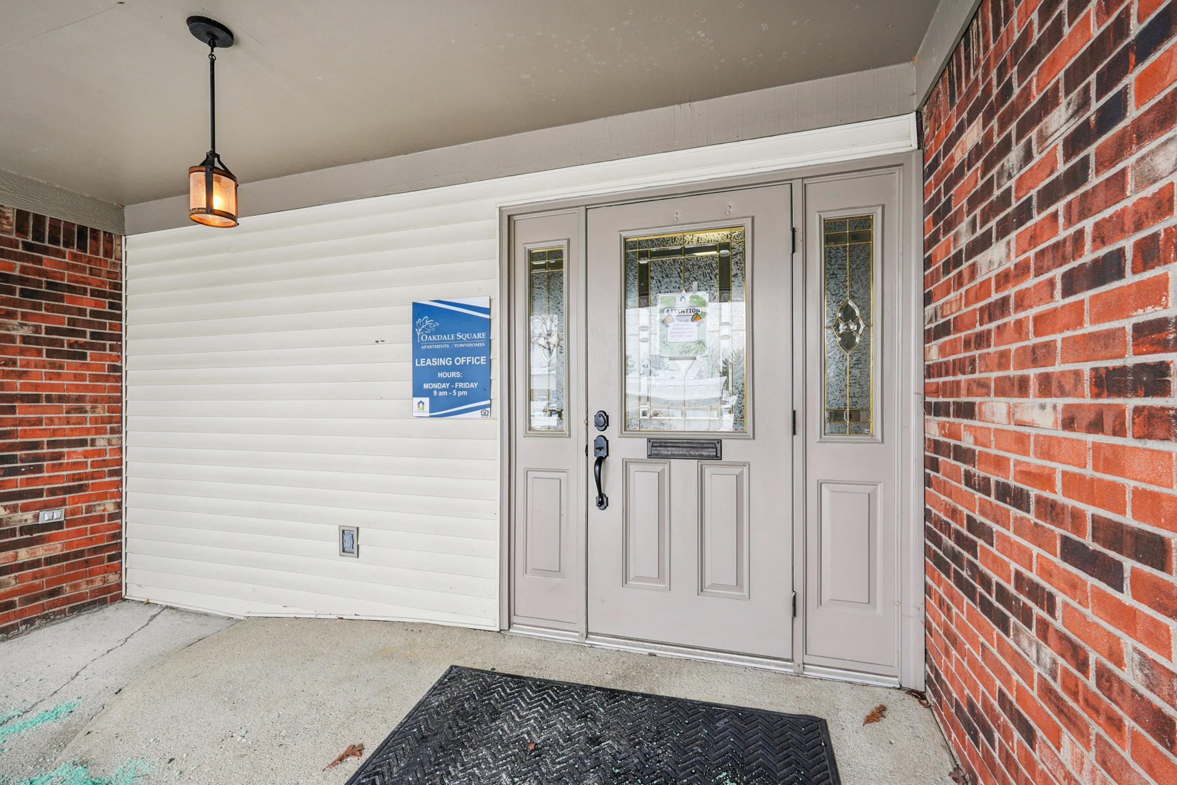 Entrance to a leasing office featuring a set of double doors with decorative glass panels, a hanging light fixture, and a sign on the wall. The area has brick and siding walls, along with a welcome mat on a concrete floor.