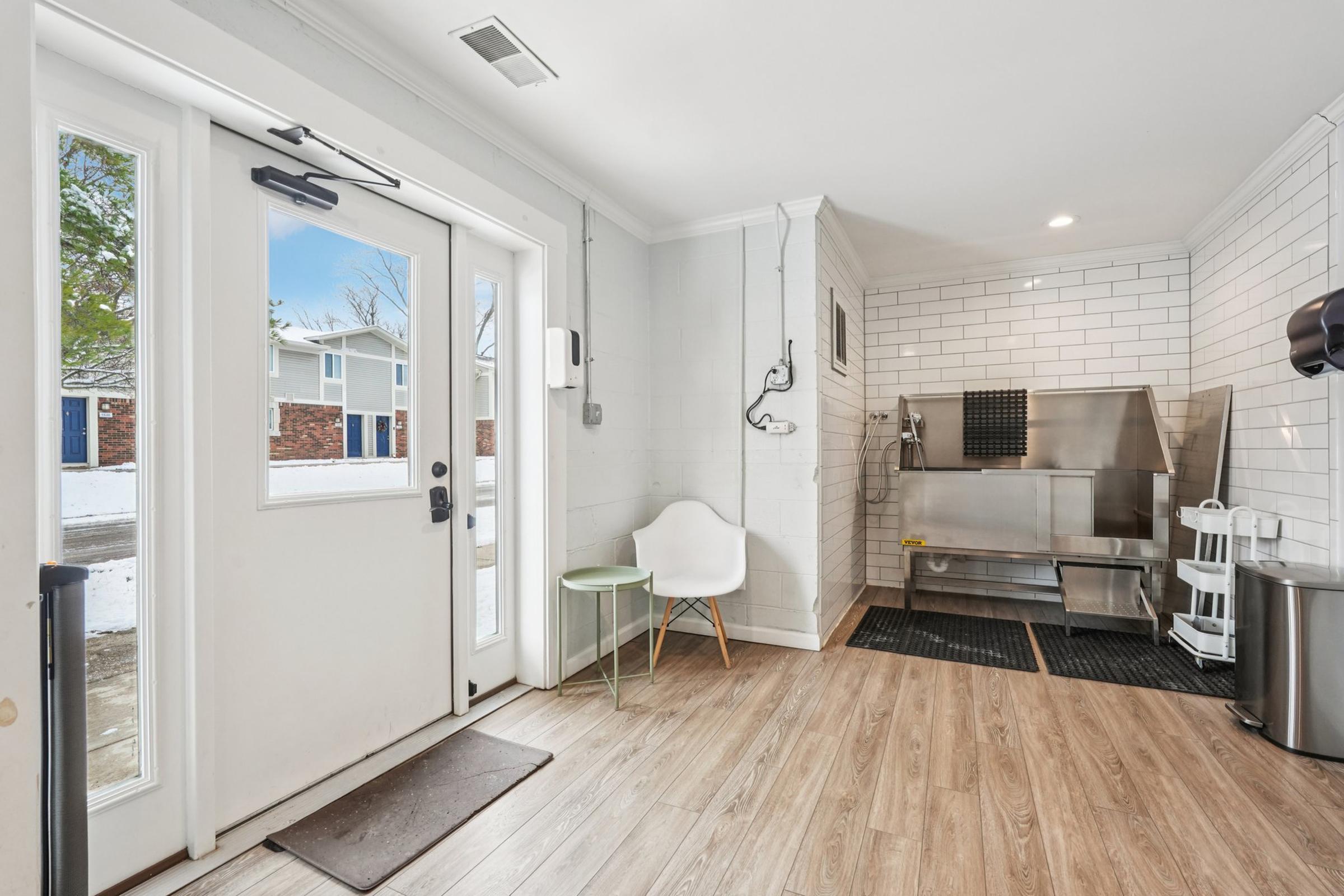 A well-lit entryway featuring a door with large windows, a small round table with a chair, and a modern kitchen area in the background. The kitchen includes stainless steel appliances and white tiled walls, with a black rubber mat on the floor, creating a clean and inviting space.