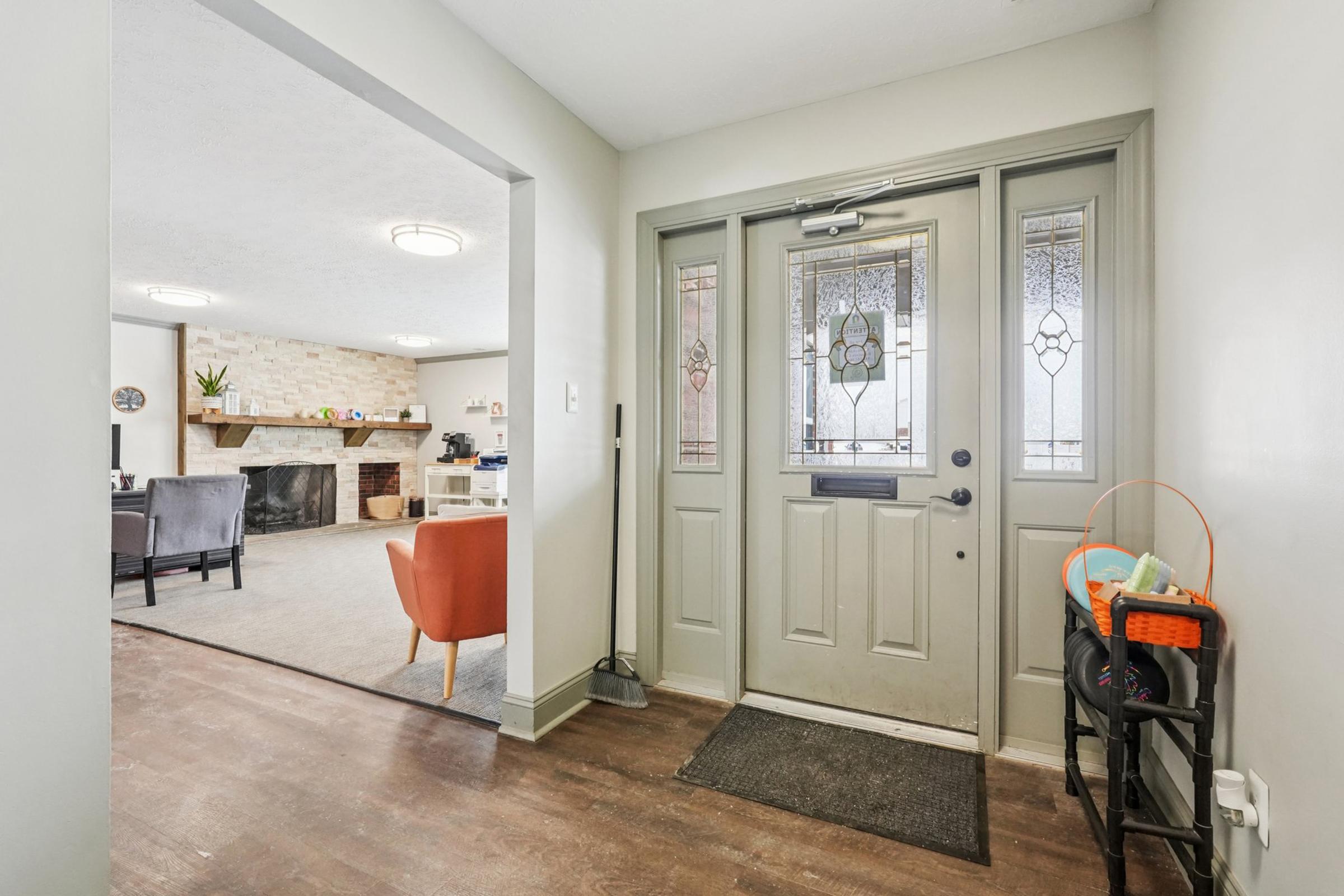 A welcoming entryway with a double door featuring decorative glass panels. To the right, there's a small table with a colorful basket. The interior has light-colored walls and wooden flooring, opening up to a cozy living area in the background, showcasing a fireplace and comfortable seating.