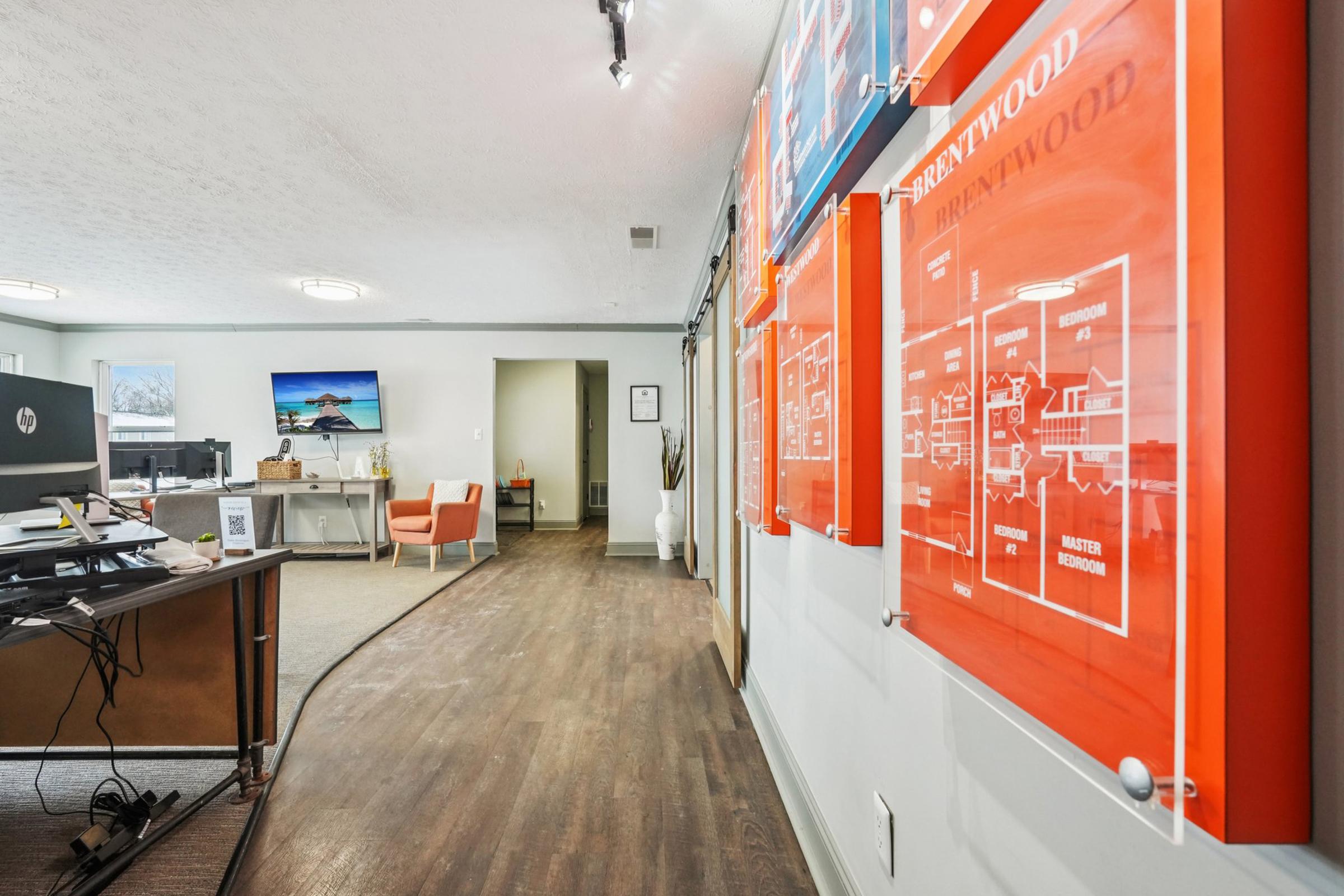 A well-lit office hallway featuring orange information boards displaying floor plans on the left. To the right, a desk with a computer is visible, along with modern seating and a decorative wall art piece in the background. The flooring is wood, contributing to a professional atmosphere.