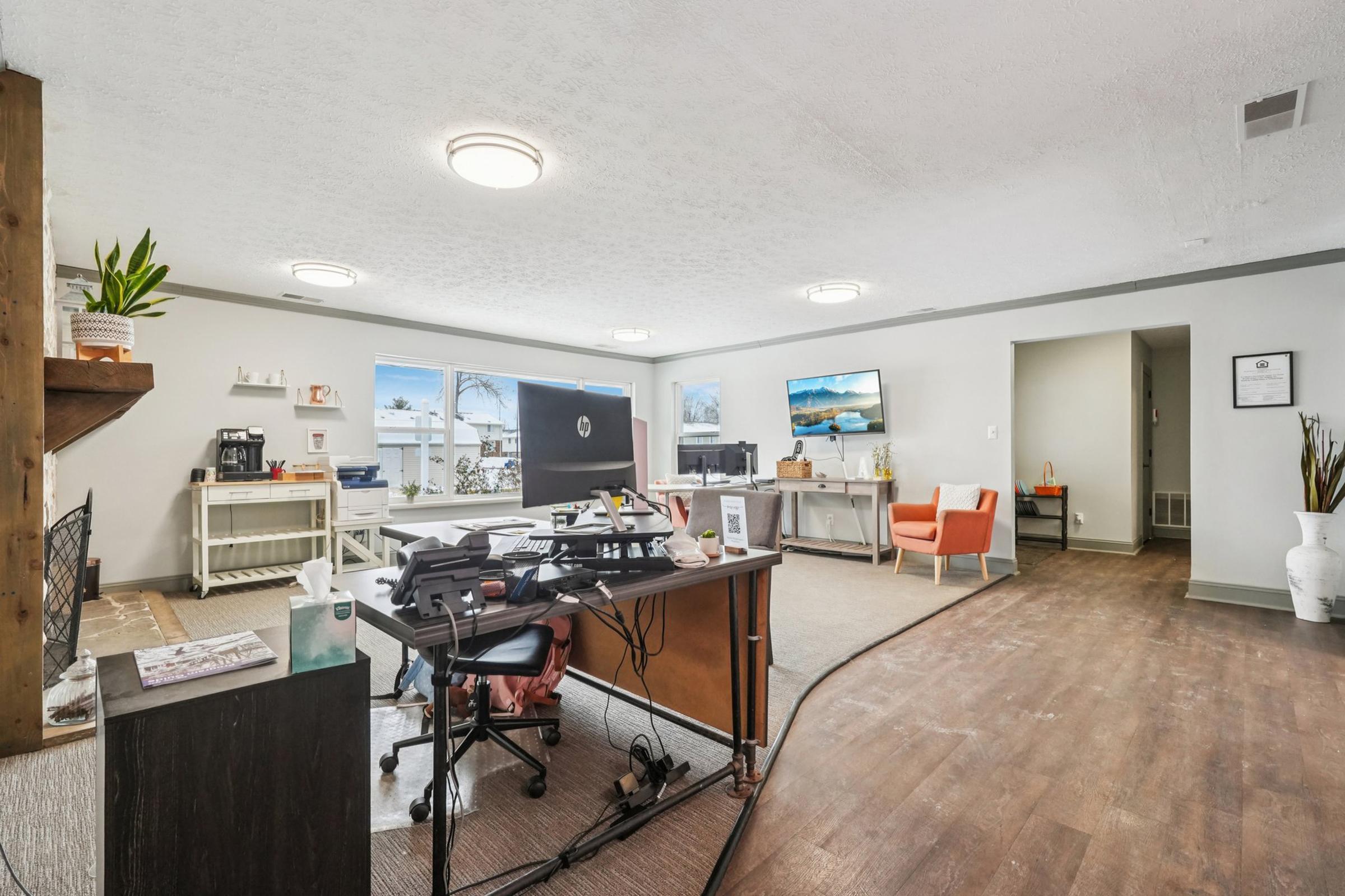 A modern office space featuring a large desk with a computer, chair, and office supplies. The room has two large windows allowing natural light, with a cozy orange chair in the corner. There are plants, light fixtures on the ceiling, and a TV displaying scenic images on the wall. The flooring is wooden.