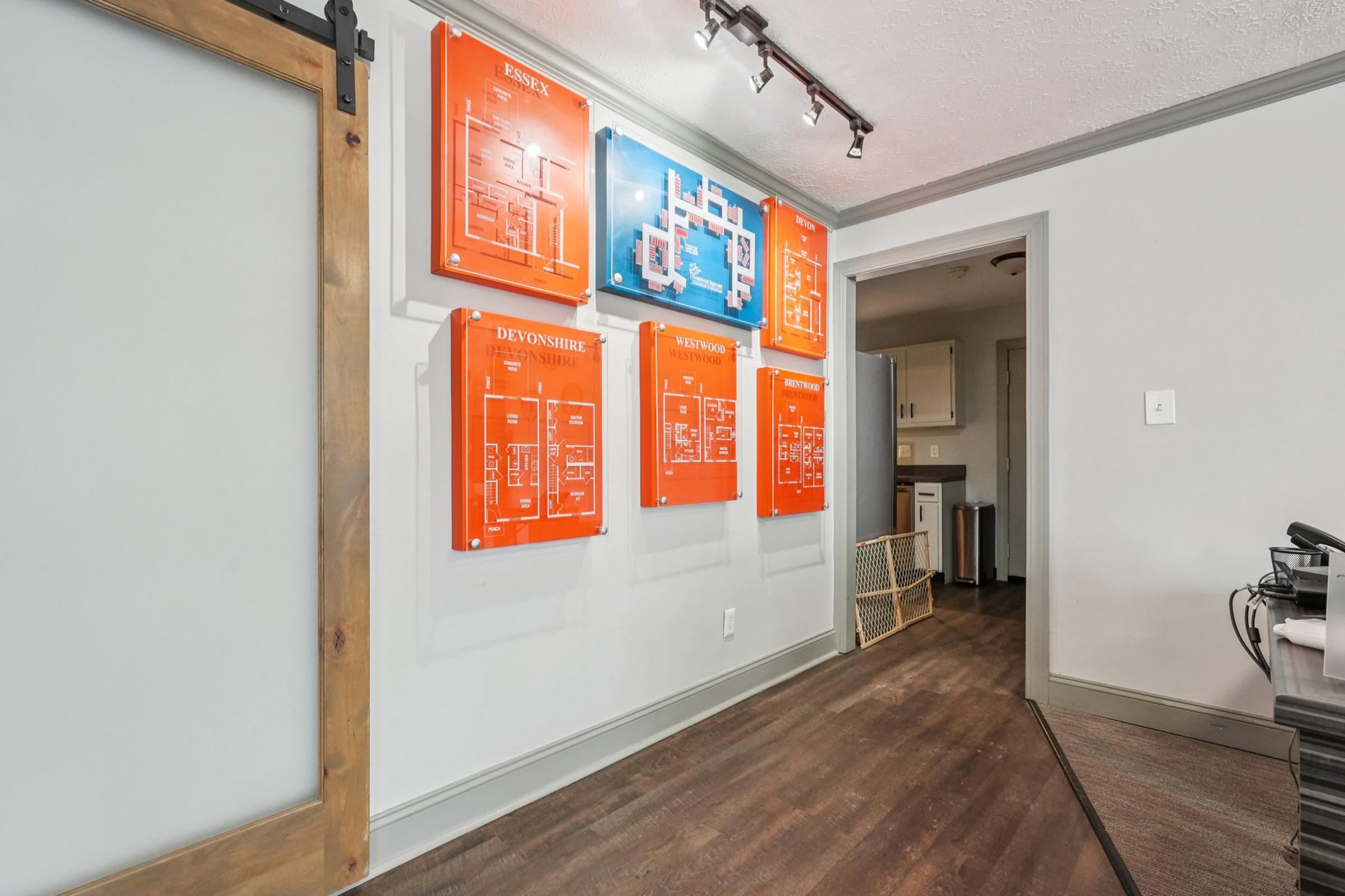 A hallway displaying multiple orange and blue floor plan diagrams mounted on the wall. The wall features a sliding wooden door on the left, leading to another room. The flooring is wooden with a modern aesthetic, and there is a glimpse of a small room or kitchen area in the background.
