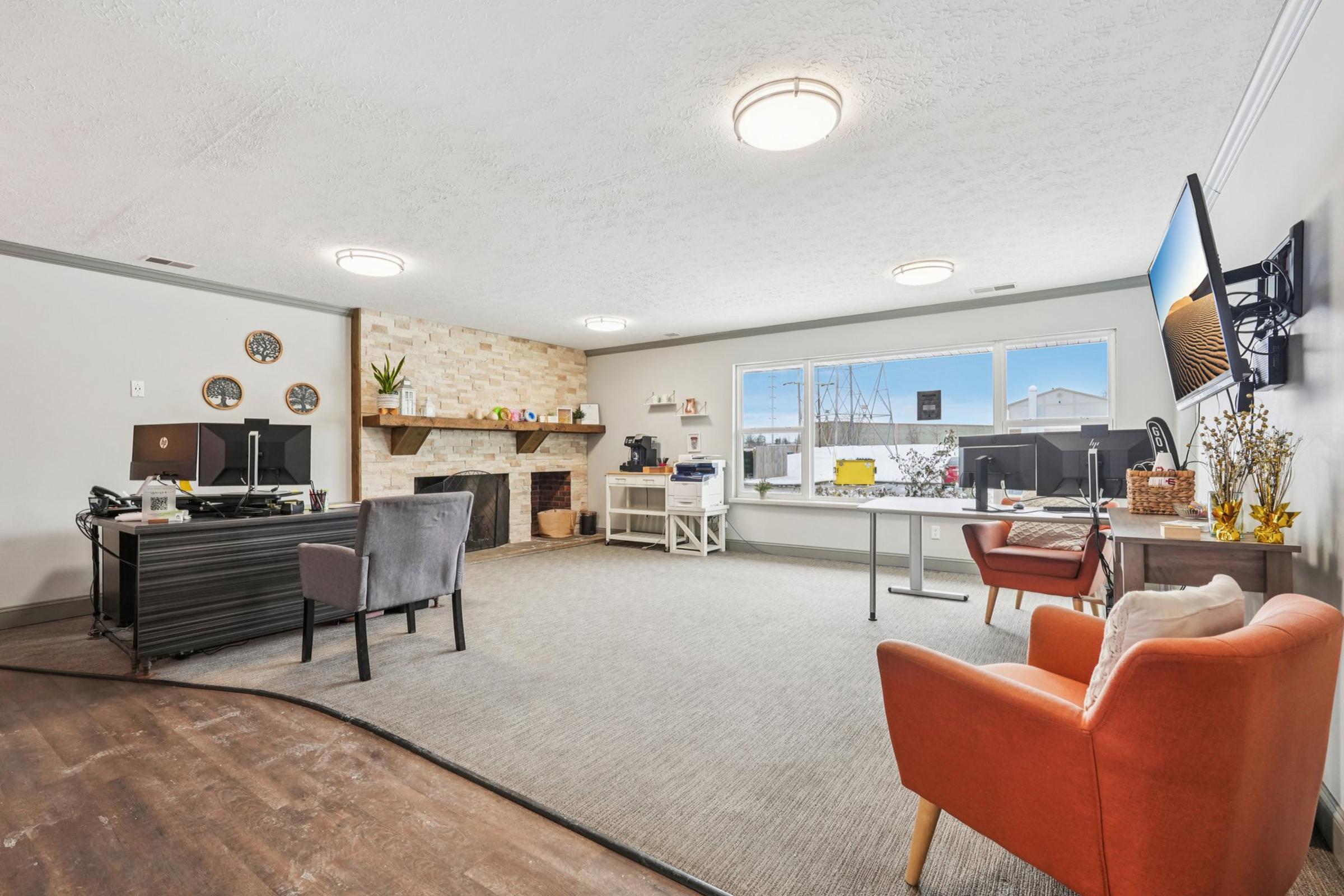 A spacious, modern office interior featuring a brick fireplace, large windows, and natural light. There are two desks with computers, a comfortable gray armchair, and an orange accent chair. Decorative items are placed on shelves, creating a welcoming workspace.