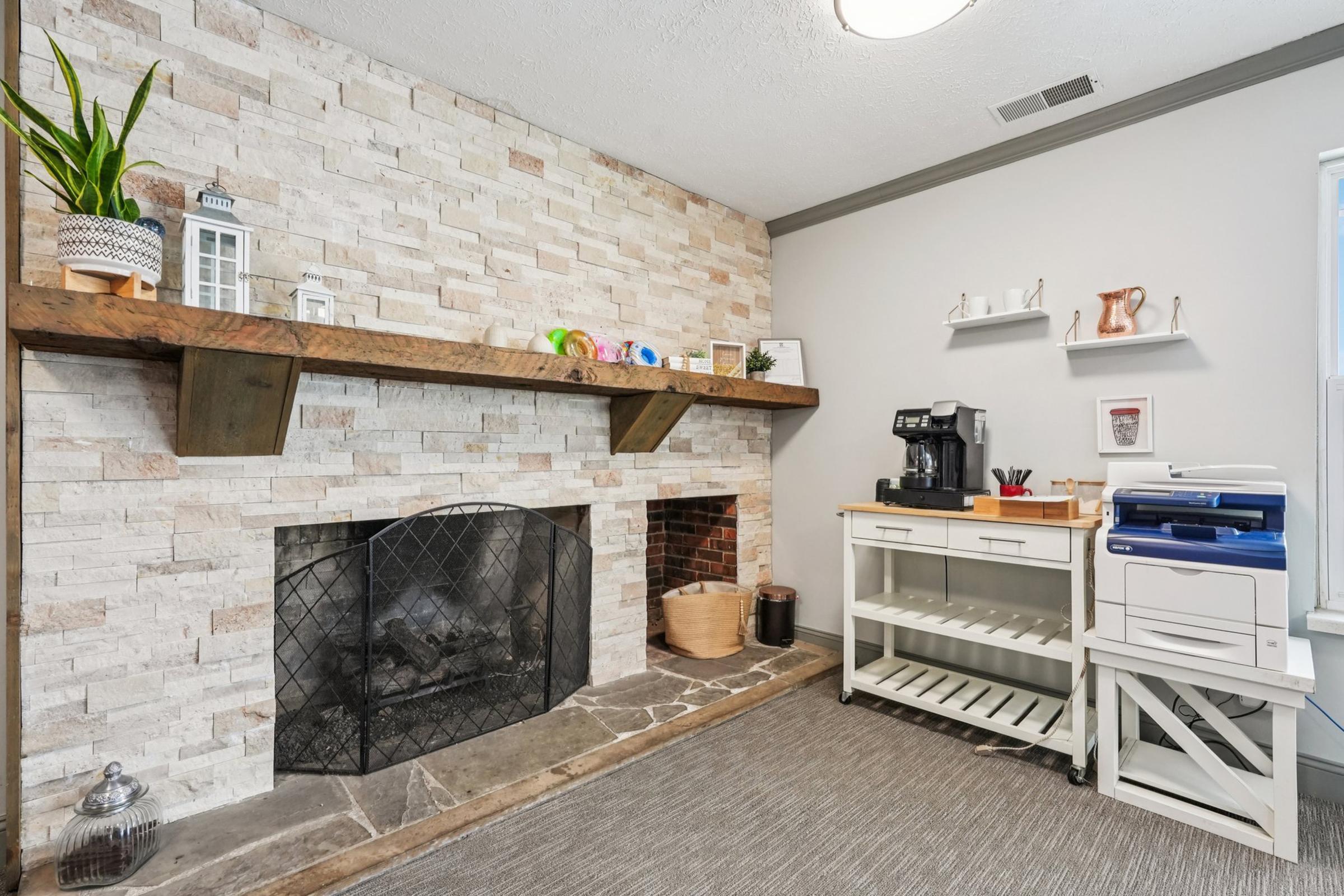 A cozy living room featuring a stone fireplace with a wooden mantel. On the mantel, there are decorative items including a lantern and plants. To the side, there's a coffee station with a coffee maker, and a white shelving unit holds a printer and decorative accents. The floor is covered with a neutral-toned rug.