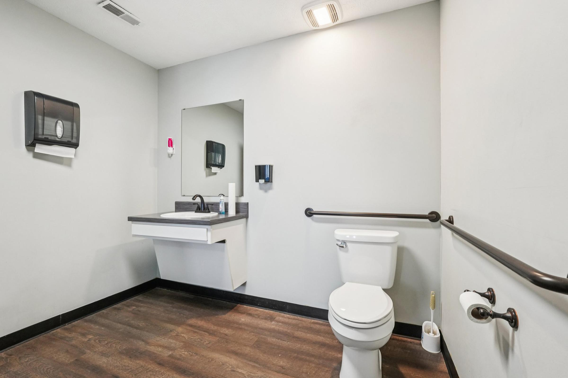 A clean and modern bathroom featuring a white toilet, a sink with a mirror above, and accessible grab bars. The walls are painted grey, and there is a wall-mounted paper towel dispenser. The floor has dark wood-like tiles, creating a contemporary and functional space.