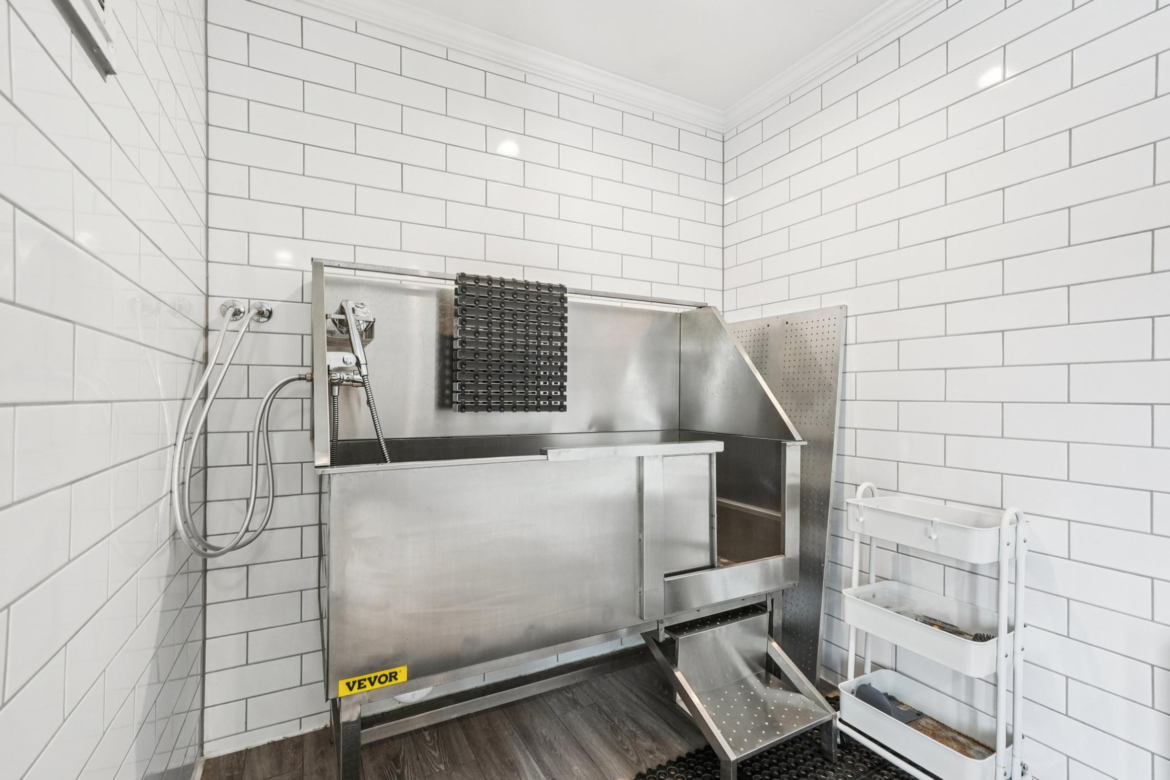 A spacious, modern dog washing station featuring a stainless steel tub with a showerhead, surrounded by white subway tiles. The area has a non-slip mat on the floor and a wheeled cart for supplies, creating a clean and functional environment for pet care.