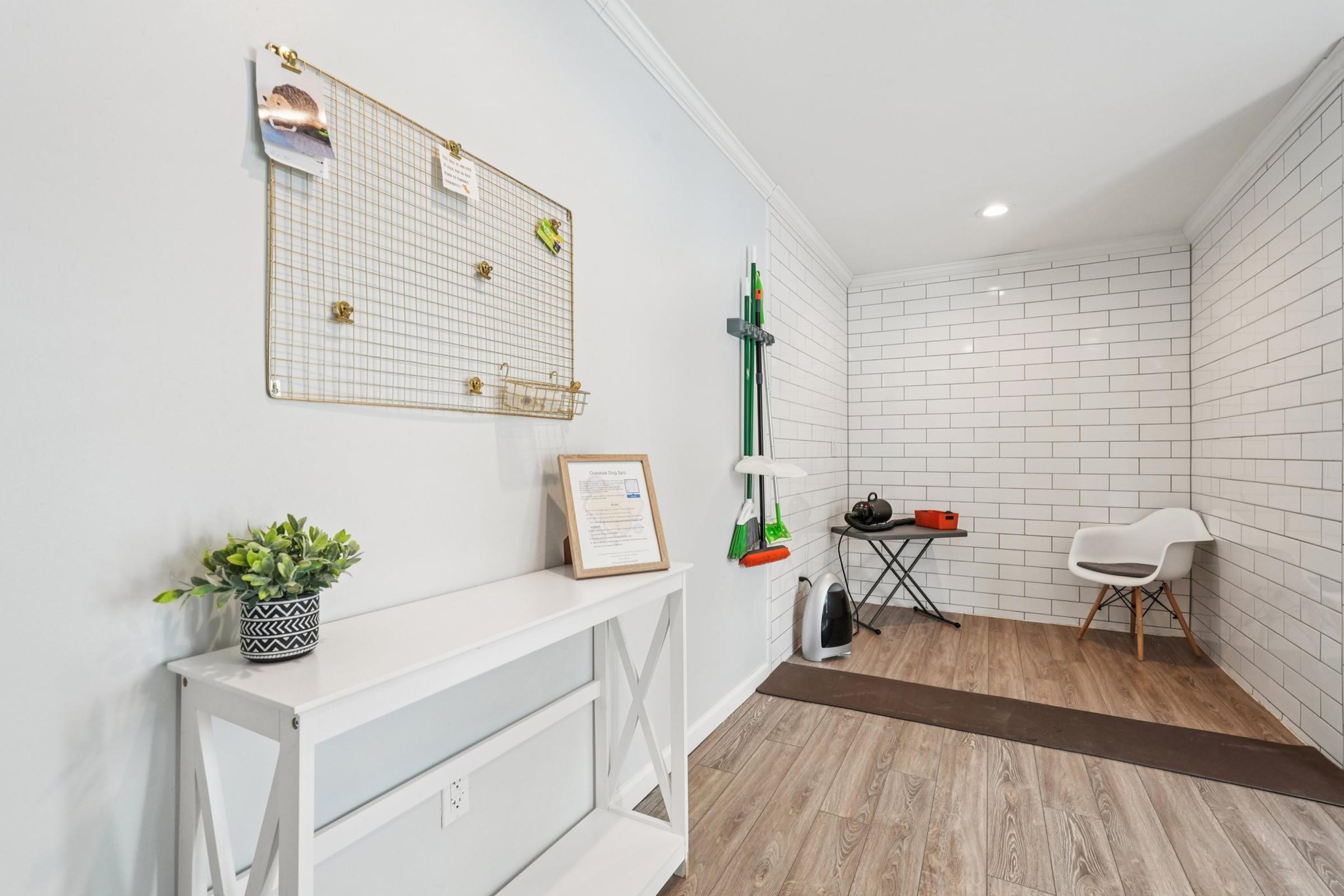 A modern entryway featuring a white wall with a wire grid board holding note cards, a small potted plant on a white console table, and a black folding table with fitness equipment. The space has light wooden flooring and white tiled walls, creating a clean and organized ambiance.