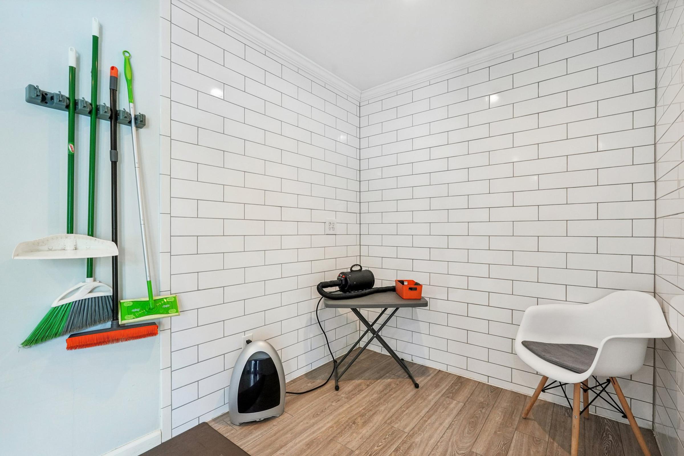 A clean corner of a room featuring white tiled walls. On the left, cleaning tools are hung on a wall-mounted rack, including a broom and a mop. In the center, a small table supports an appliance and a black vacuum cleaner. A modern white chair with wooden legs is positioned next to the table.