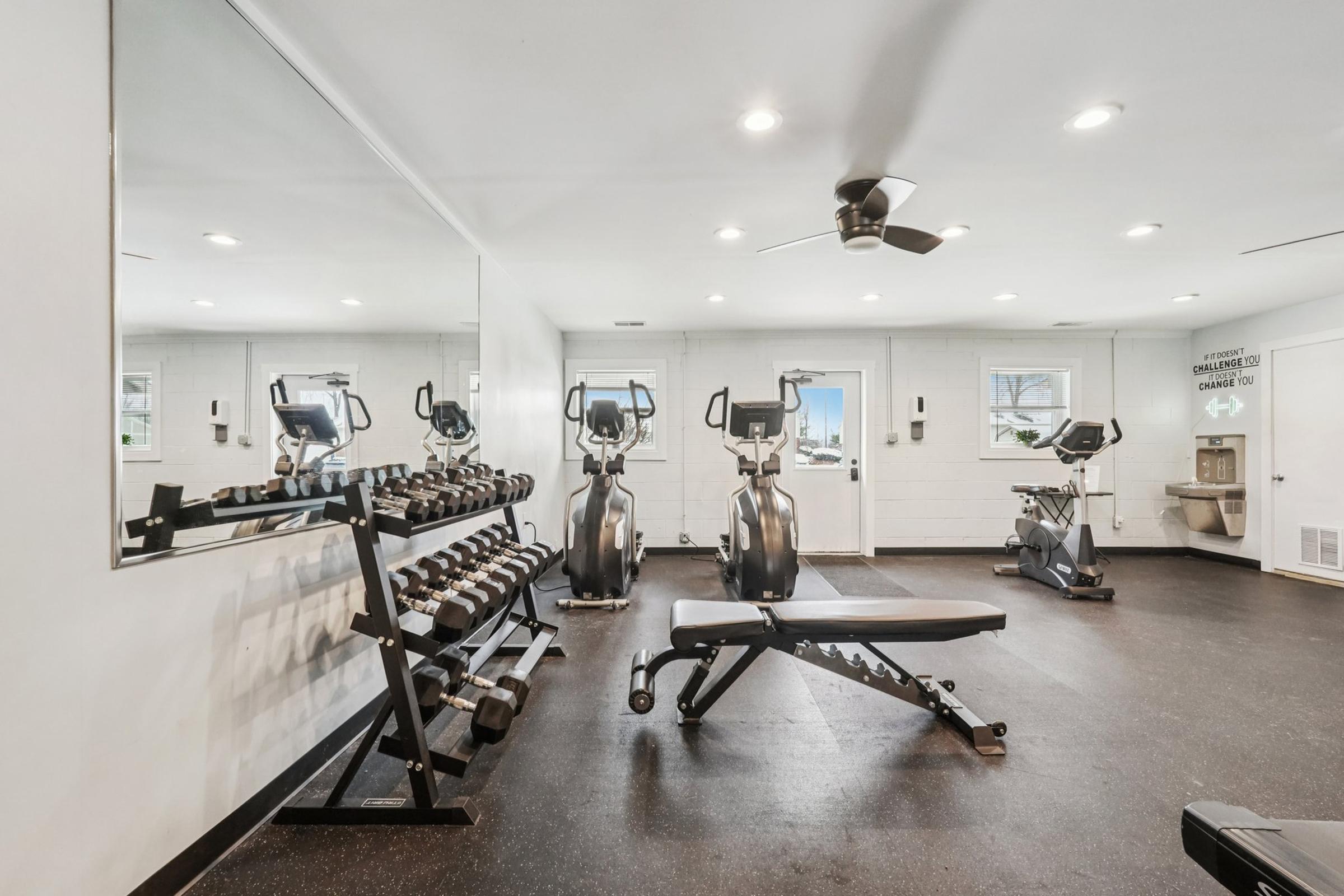 A well-lit fitness room featuring a mirror, a set of dumbbells on a rack, a weight bench, and two elliptical machines. The space is equipped with a ceiling fan and has large windows providing natural light. The walls are painted white, and the flooring is dark rubber.