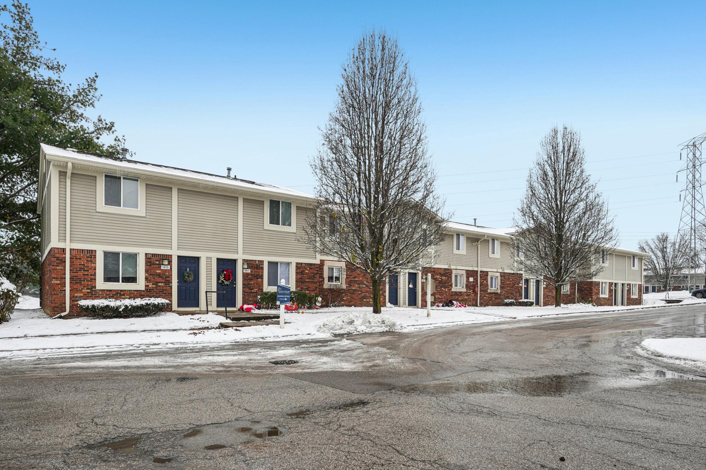 A residential street lined with several two-story townhouses. Each unit features a front door decorated with a wreath and surrounded by snow. The area is wintery with patches of snow on the ground, and there's a clear blue sky in the background.