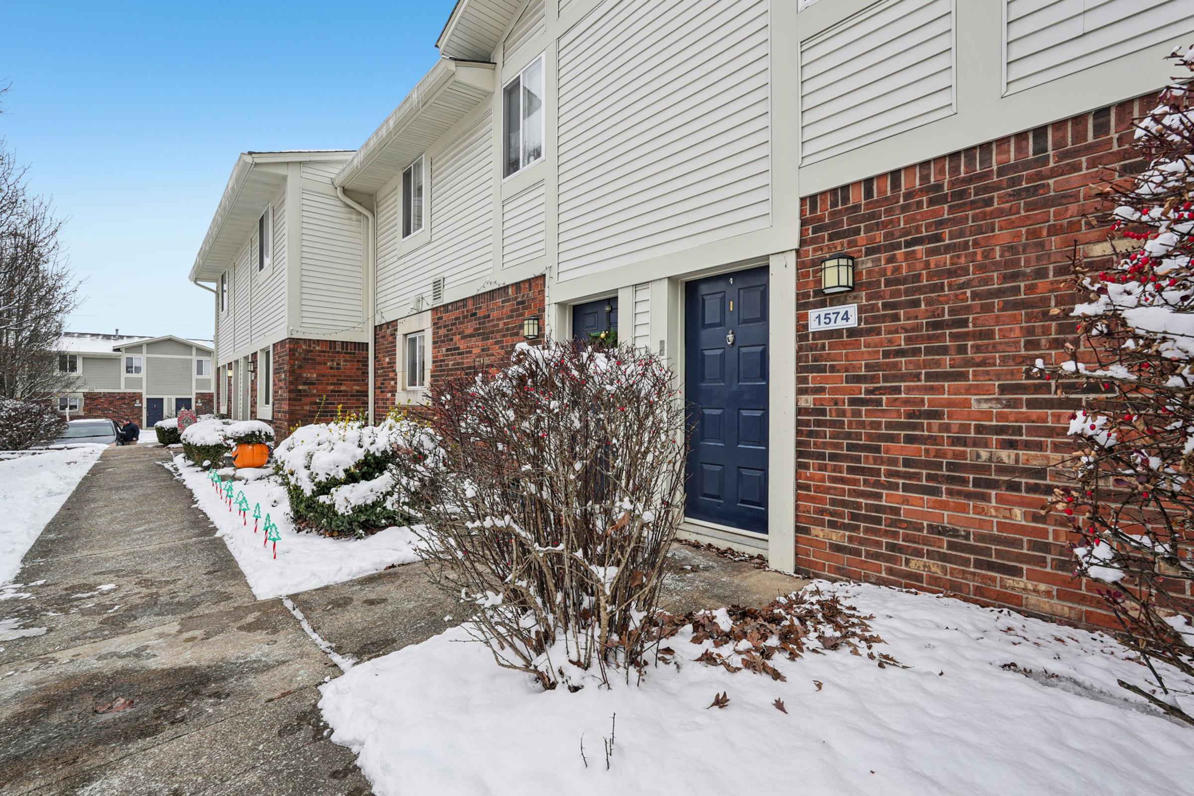 A residential building with brick and siding exterior. A front walkway leads to a blue door marked with the number 1574. Snow covers the ground and bushes, while festive lights line the walkway. A pumpkin decoration is visible on the left side of the entrance, contrasting with the winter scene.