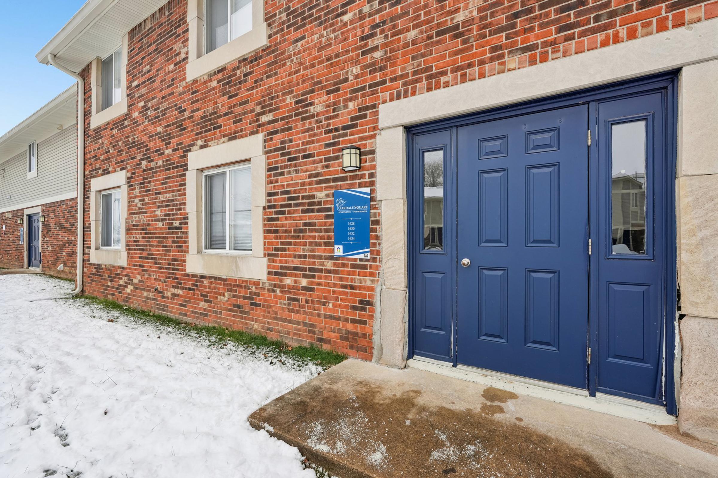 A brick building with a blue double door entrance. The door has two small windows and a light fixture above it. Next to the door is a sign attached to the wall. Snow is visible on the ground in front of the entrance. The area is clear of obstructions, and the building's exterior shows a well-maintained appearance.