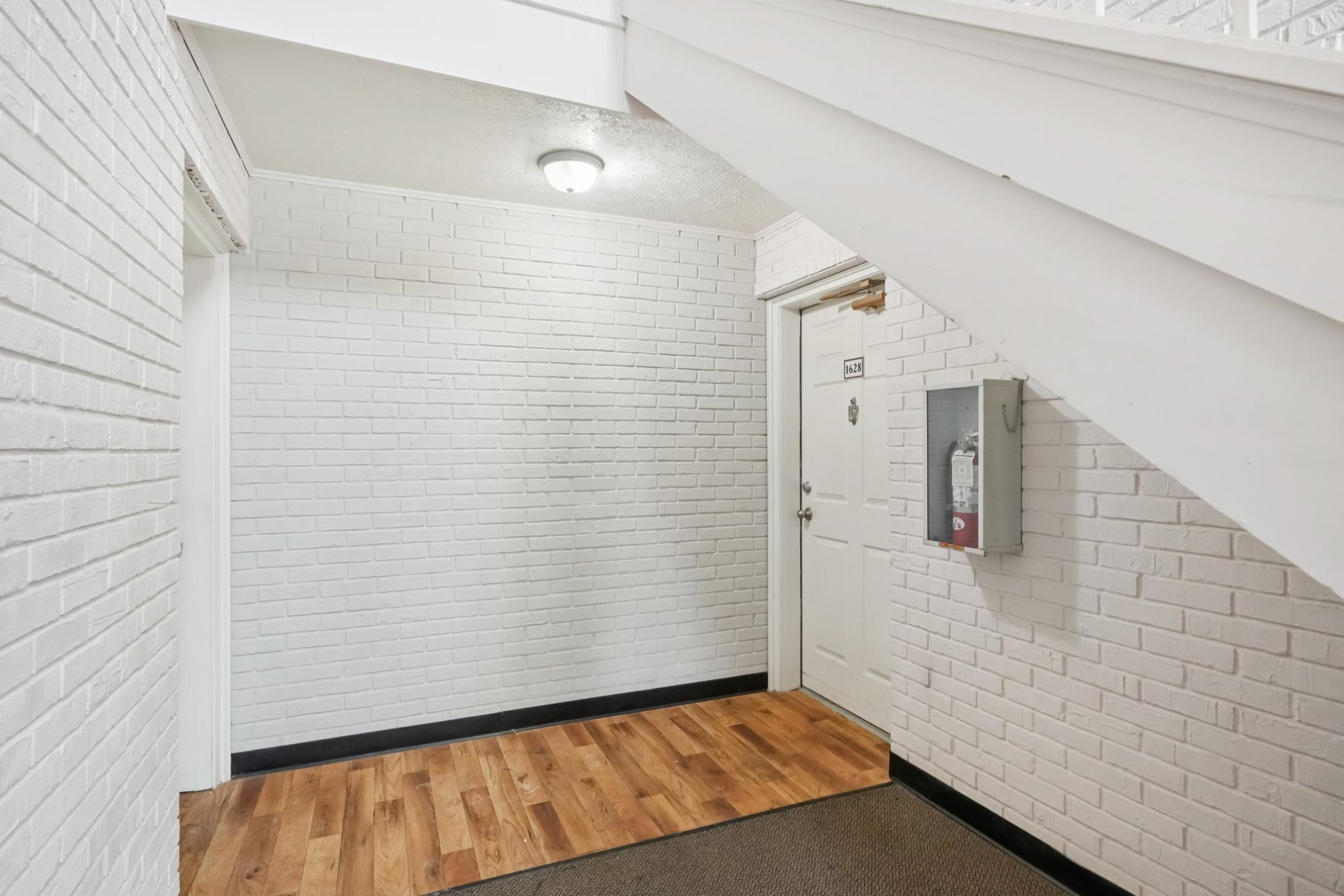 Interior hallway featuring white brick walls and wooden flooring. A door is visible on the right side, with a fire extinguisher mounted on the wall next to it. The staircase is located at the top left, and the area is well-lit with overhead lighting.