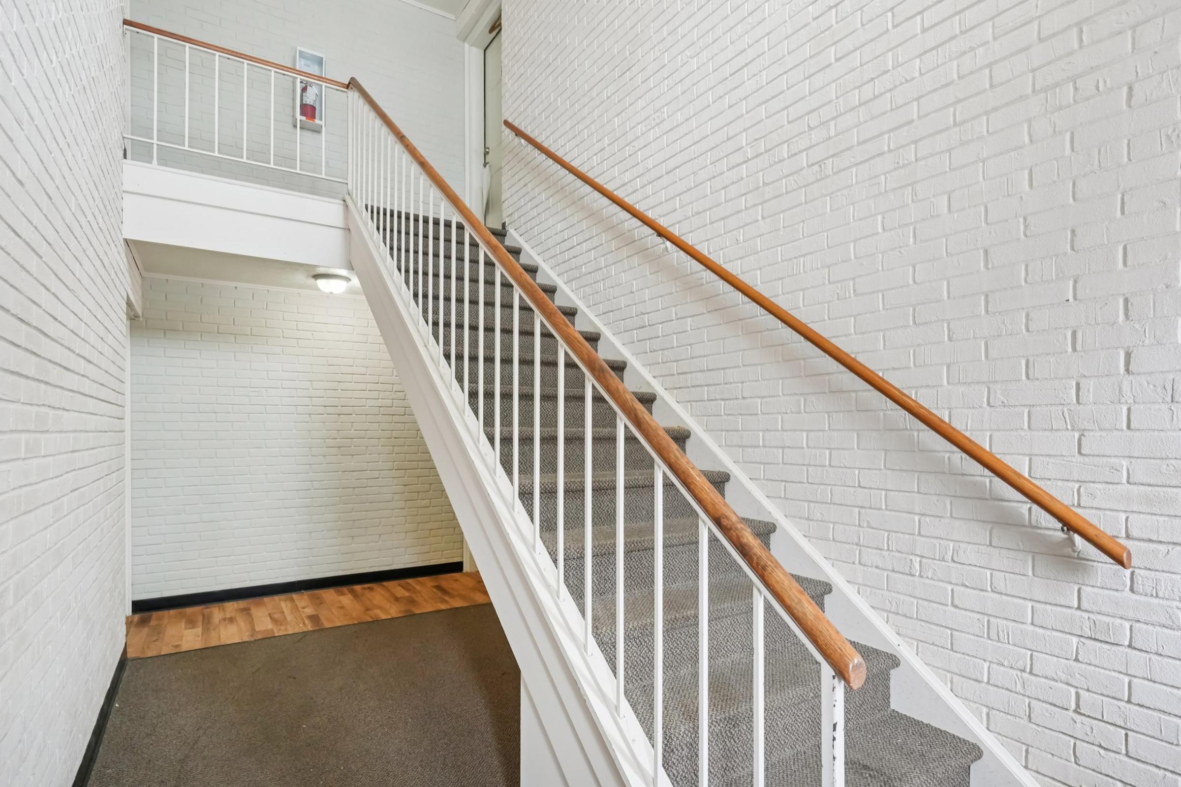 A well-lit staircase in a building with white brick walls and a wooden handrail. The stairs are carpeted, and the landing area features a wooden floor. There is a door at the top of the stairs, and the overall ambiance is clean and modern.