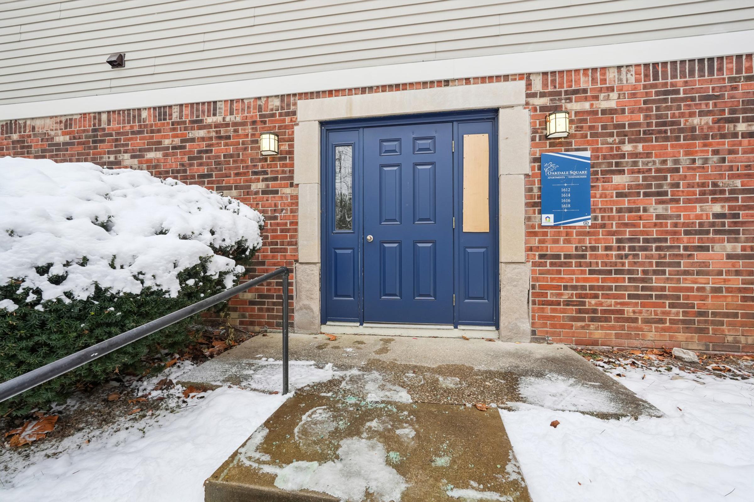 A blue front door with a boarded-up window, set in a brick wall, is surrounded by snow-covered bushes. A set of stairs leads up to the door, and there is a sign next to the entrance. The sidewalk is partially cleared of snow, indicating recent winter weather.
