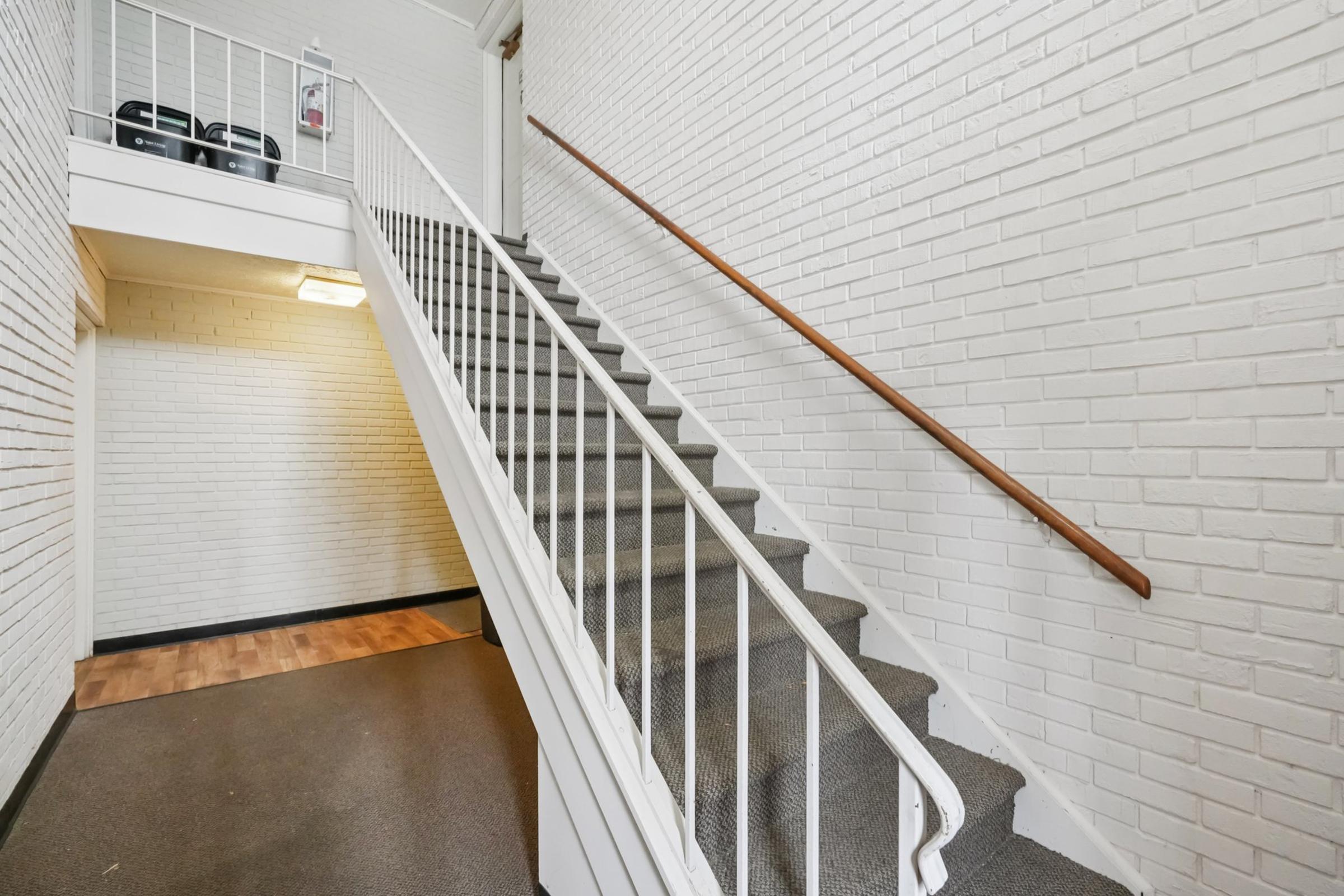 A well-lit staircase with white brick walls, featuring a wooden railing and gray carpeted steps. The top landing is partially visible, and a small area with two black objects is located near the ceiling. The floor is finished with a light-colored wood.