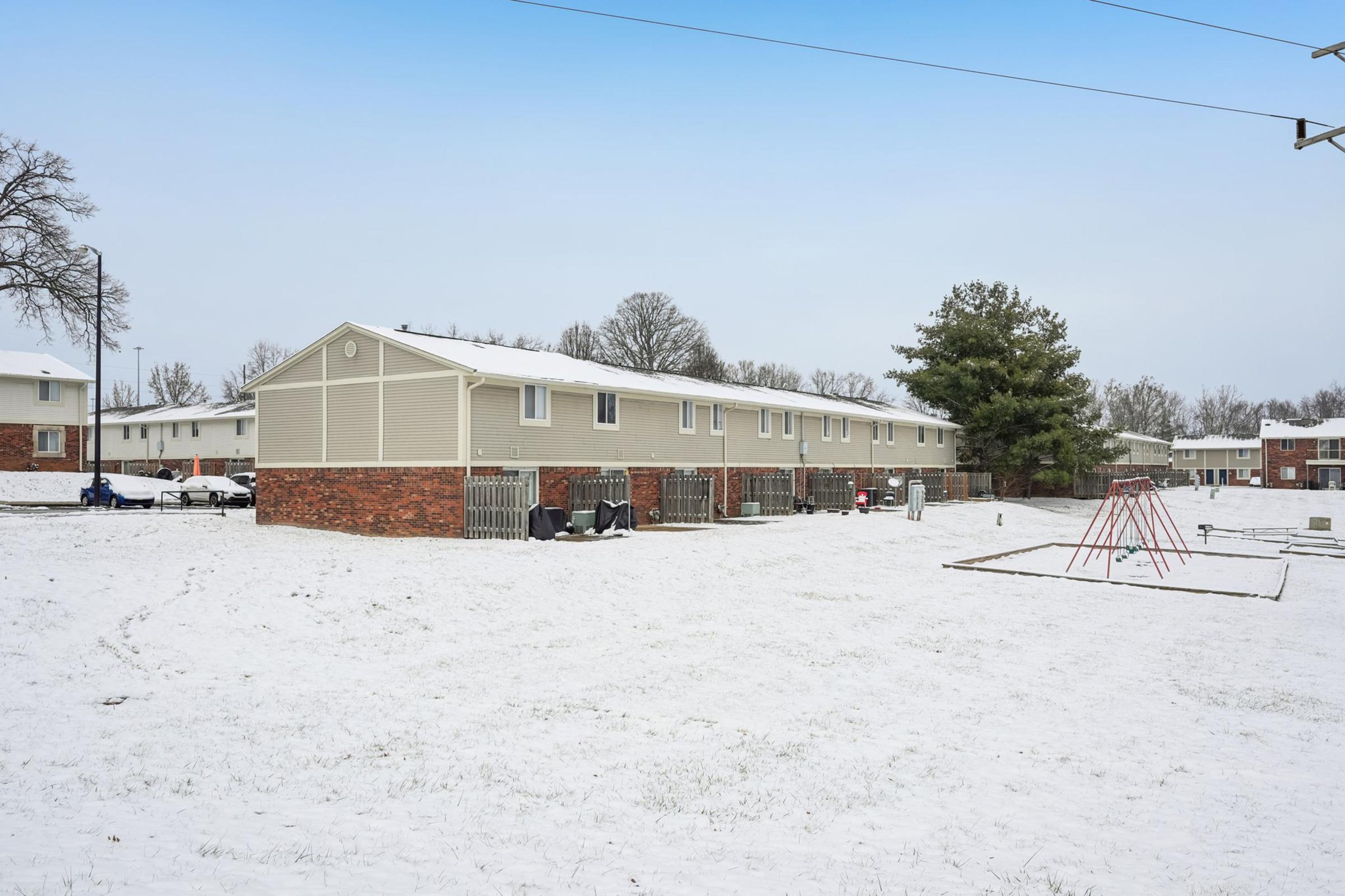 A winter scene featuring a row of apartment buildings covered in snow. In the foreground, there is a grassy area blanketed with snow, and a playground with swings and slides is visible. Trees are in the background, partially bare, indicating the cold season.