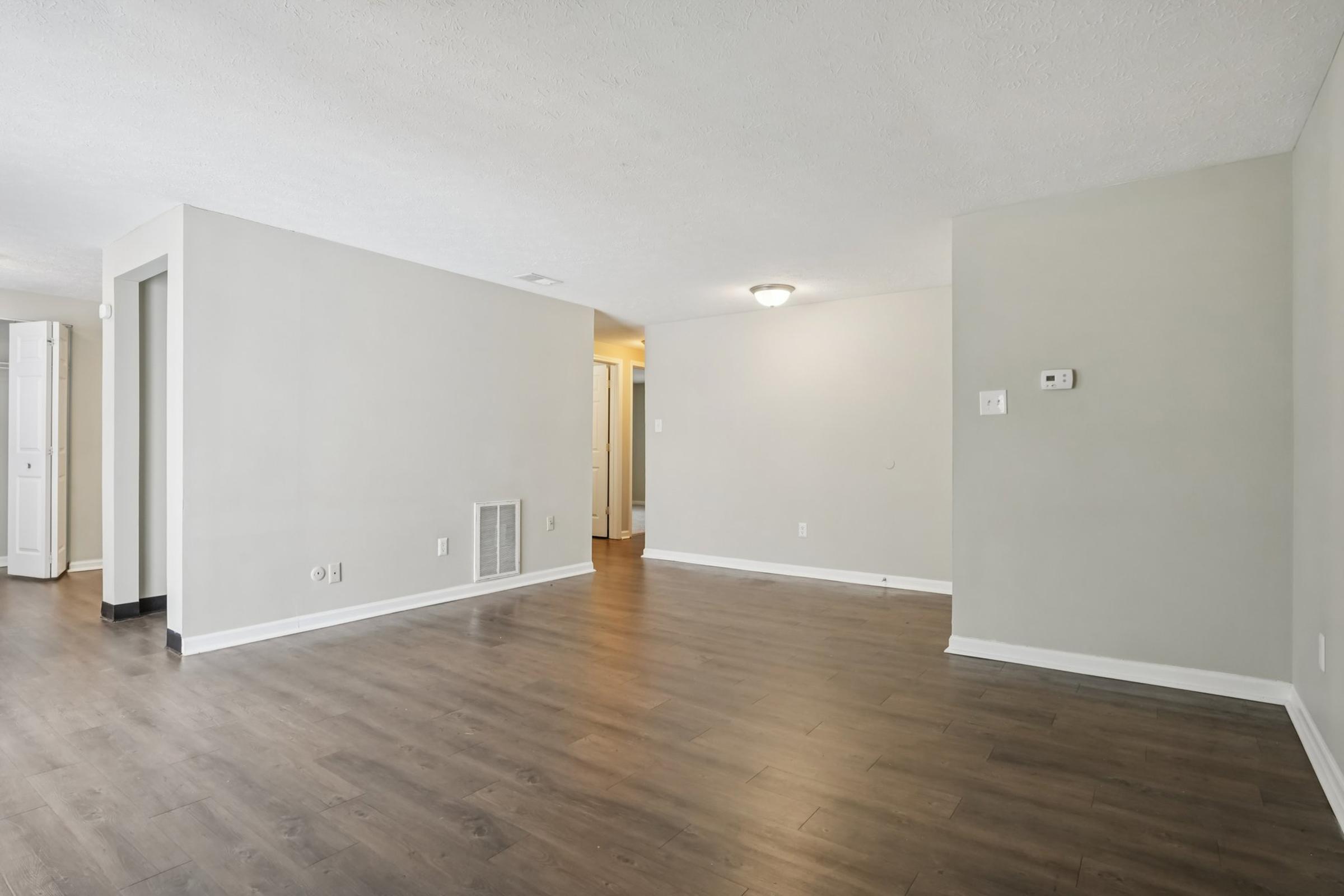 An empty room with light-colored walls and wooden flooring. There is a doorway leading to another room, a light fixture on the ceiling, and a wall-mounted thermostat. A vent is visible on one wall, and adjacent walls have minimal decorations, creating a spacious and airy atmosphere.