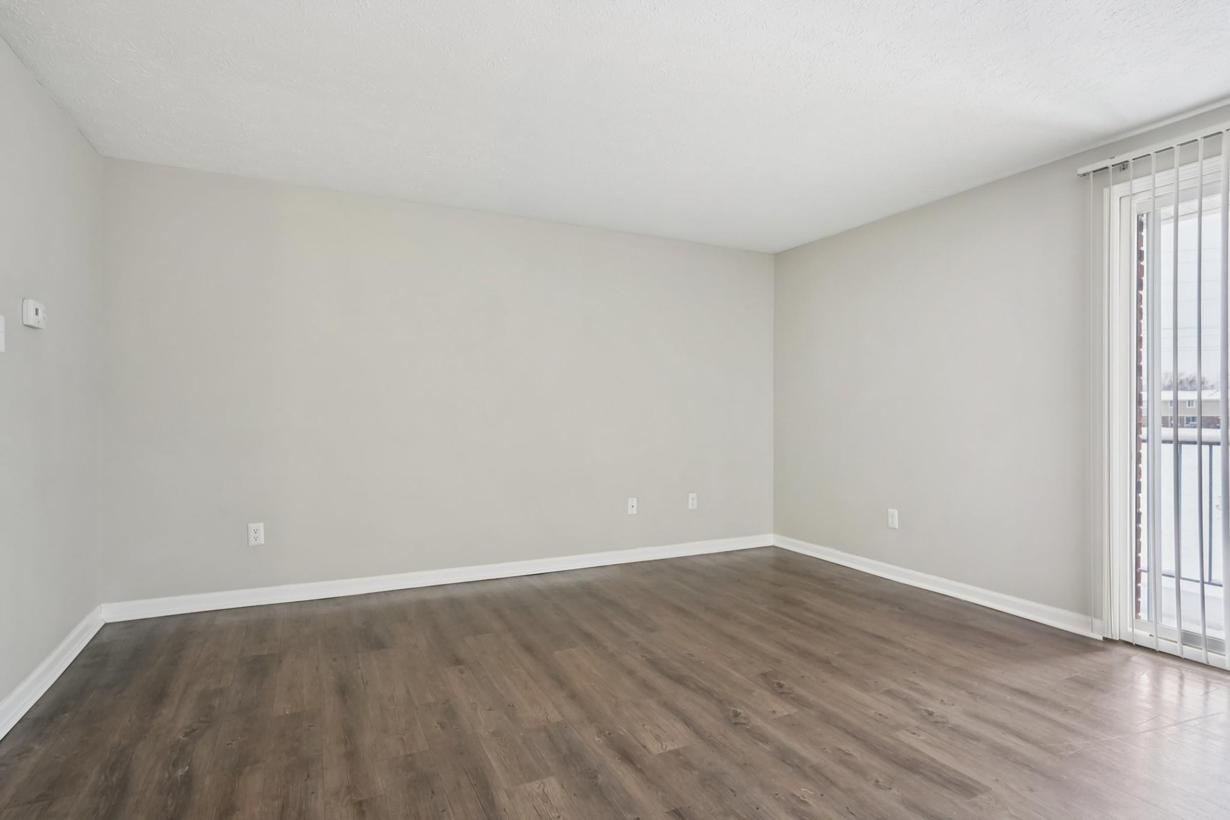 A spacious, empty room with light gray walls and dark wood laminate flooring. It features a sliding glass door that leads to a balcony, allowing natural light to enter. There are no furnishings or decorations, emphasizing the room's openness and potential for customization.