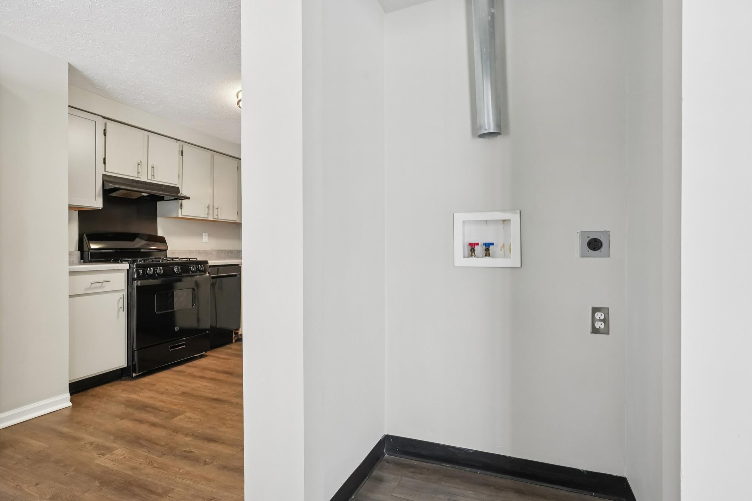 A view of a kitchen area featuring black appliances, including a stove and refrigerator, with white cabinetry. On the right, there is a small nook containing electrical outlets and a vent pipe. The floor is covered in light-colored wood laminate, and the walls are painted a neutral shade.