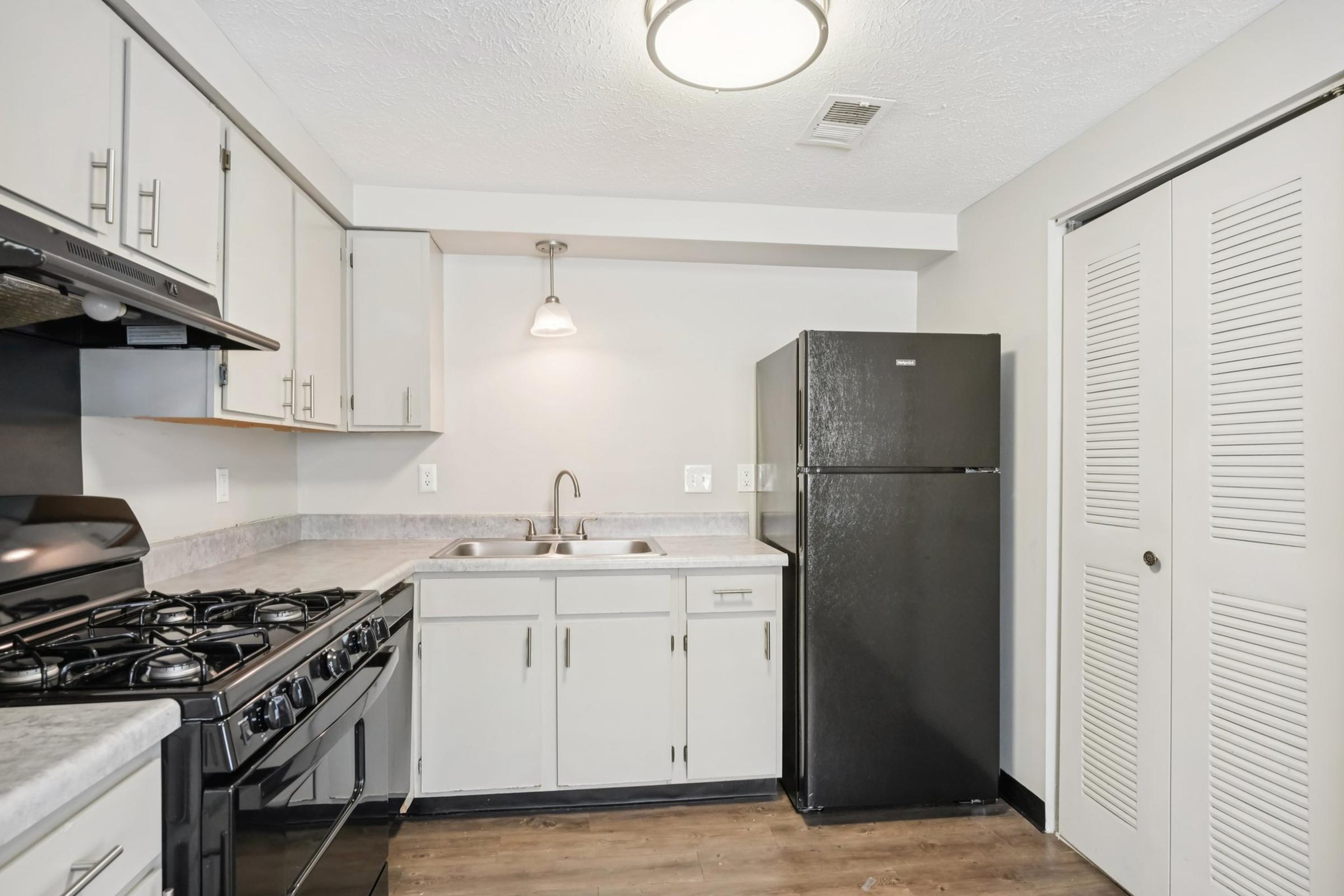 A modern kitchen featuring stainless steel appliances, including a gas stove and a black refrigerator. The countertops are light-colored, and there's a single sink under a pendant light. The cabinets are a neutral shade, and the floor is wood-like vinyl, giving the space a clean and contemporary look.