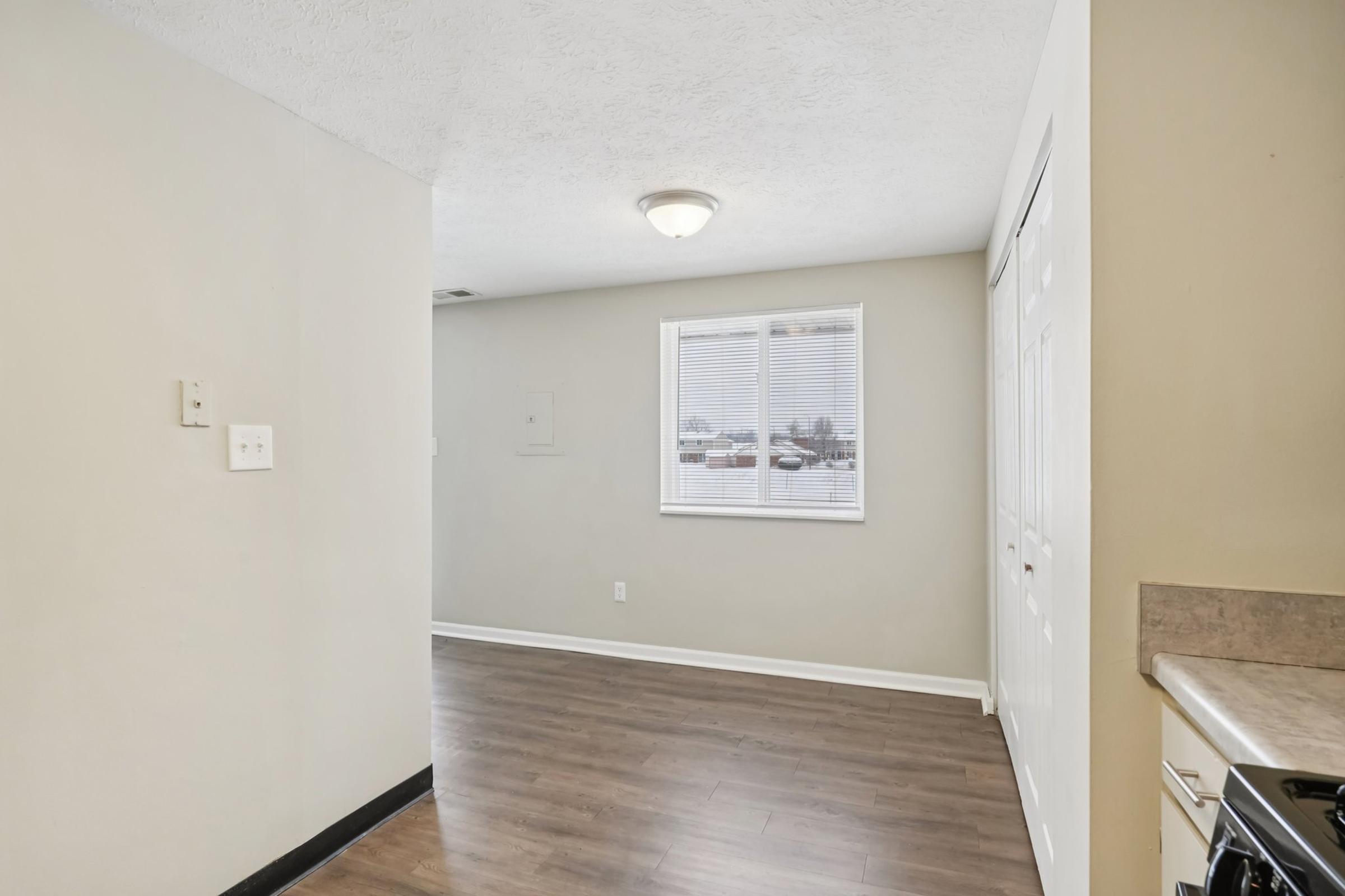 A spacious, well-lit room with light gray walls and brown laminate flooring. A window with white blinds allows natural light to enter. There's a corner with a white door and a kitchen area visible on the right side, featuring a stove. The overall atmosphere is clean and inviting.