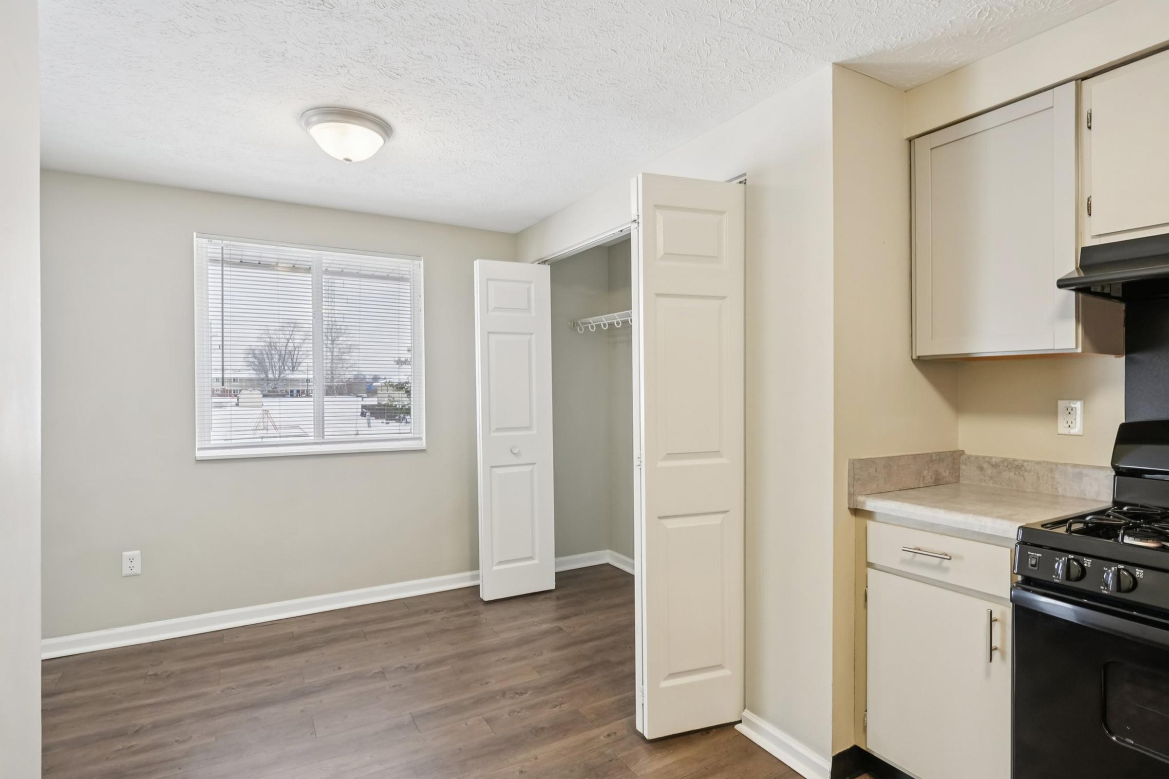 Bright, empty room with light-colored walls and wooden flooring. A window lets in natural light, and there is a closet with sliding doors. The kitchen area features a stovetop and countertop, with cabinets above and below. Overall, the space is clean and ready for furnishing.
