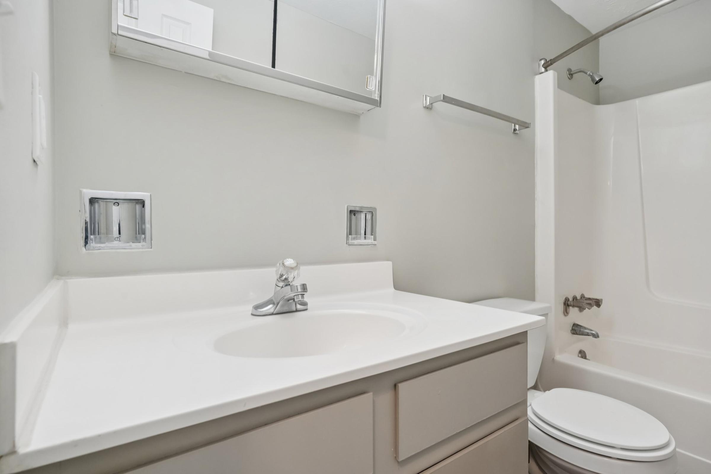 A modern bathroom featuring a white countertop with a sink, a wall-mounted mirror, and a gray wall color. The design includes basic fixtures, a shower-tub combination, and a toilet. The space is uncluttered and well-lit, providing a clean and minimalist aesthetic.