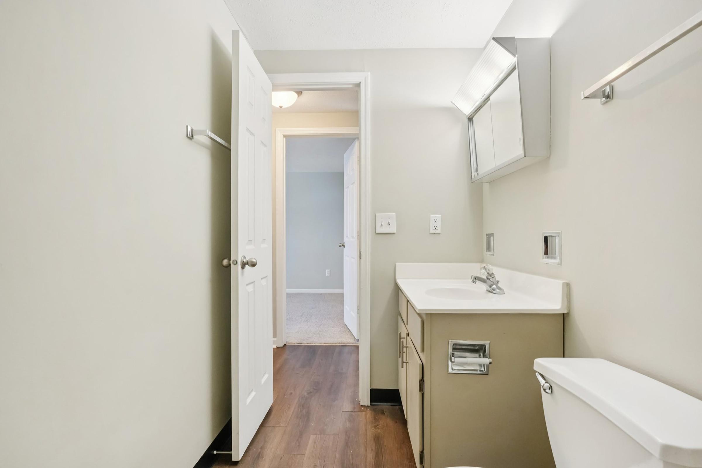 A clean, modern bathroom featuring a white sink cabinet, a mirror above the sink, and a toilet. The walls are painted a light color, and there is a door leading to another room. The flooring is dark wood, adding contrast to the light-colored walls.