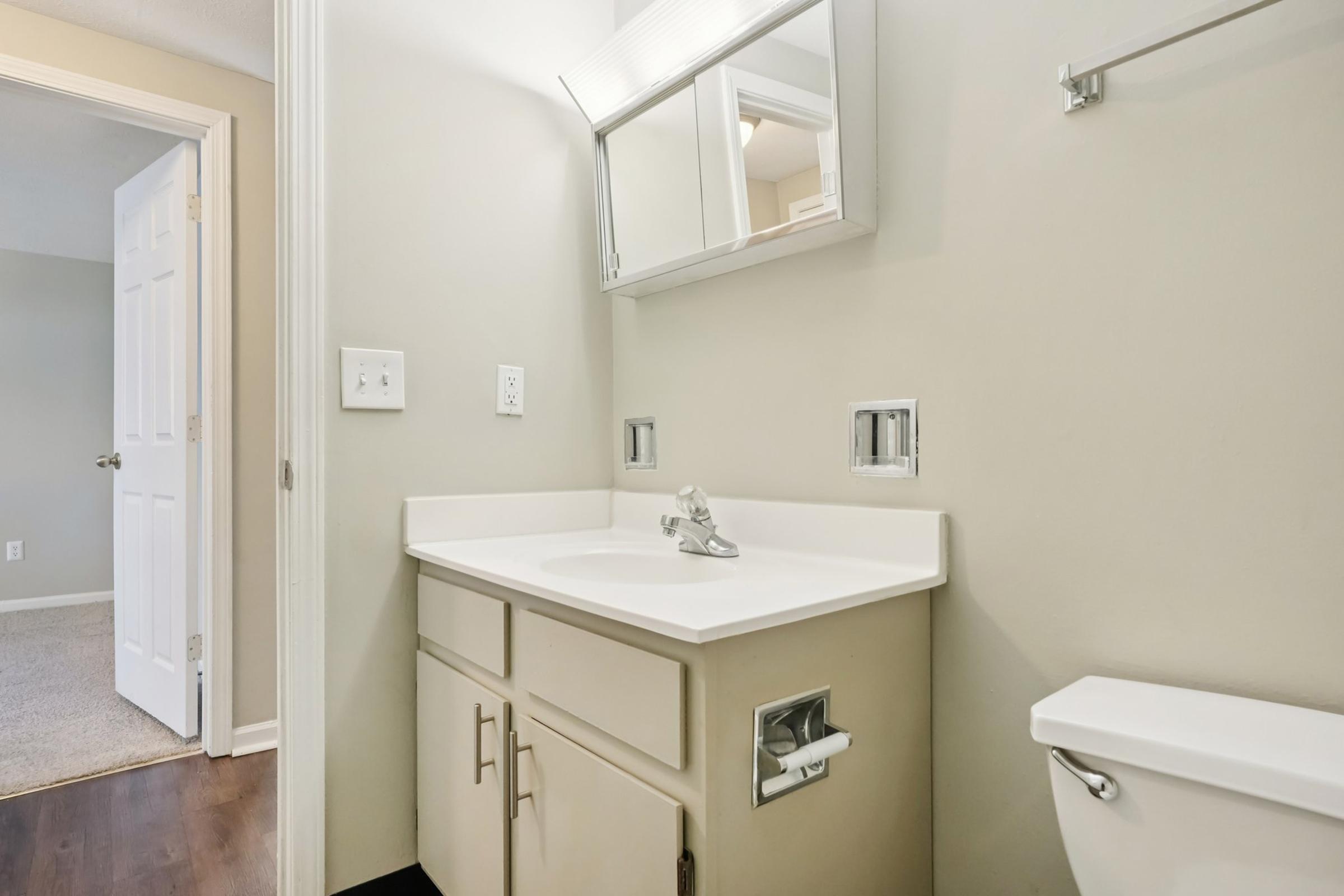 A small bathroom with a white vanity and sink, a mirror above the sink, and a toilet nearby. The walls are painted a light color, and there is a door leading to another room, partially visible in the background. The floor has dark wood or laminate tiles.