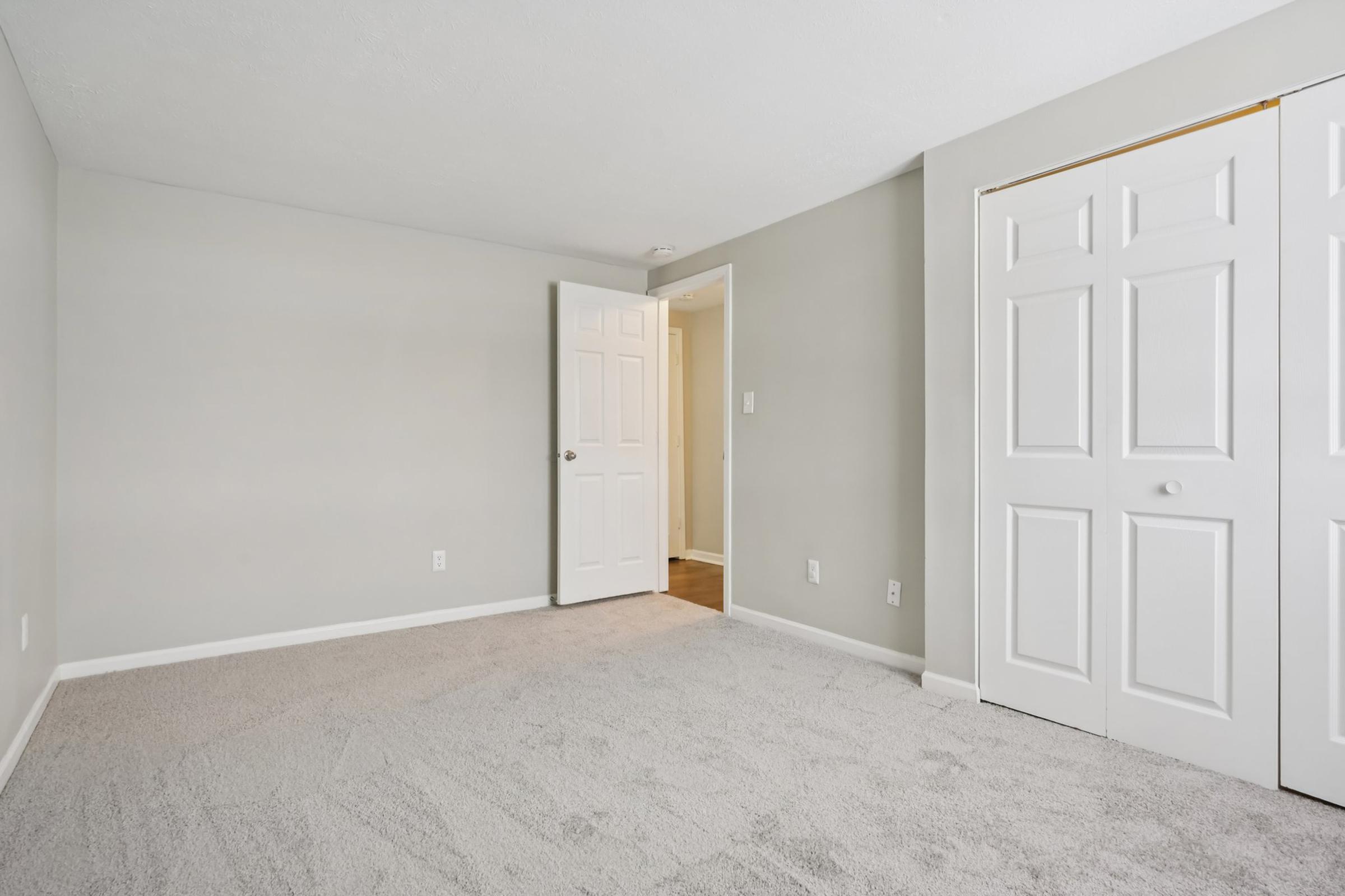 A spacious room with light gray walls and new carpeting. A closed white door leads to another room, and there is a sliding door closet on the right. The room is well-lit and appears vacant, ready for furnishing.