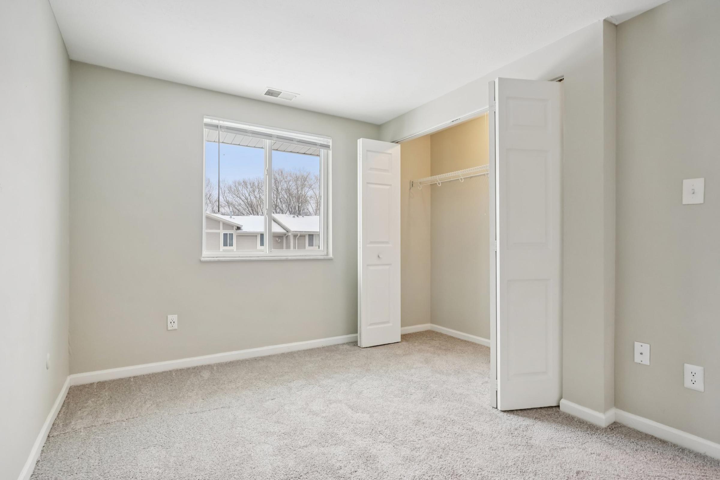 Interior of a light-colored bedroom with neutral walls and carpet. A window allows natural light to enter, and there is a closet with double doors on one side. The room appears spacious, with no furniture, creating a blank canvas for potential arrangement.