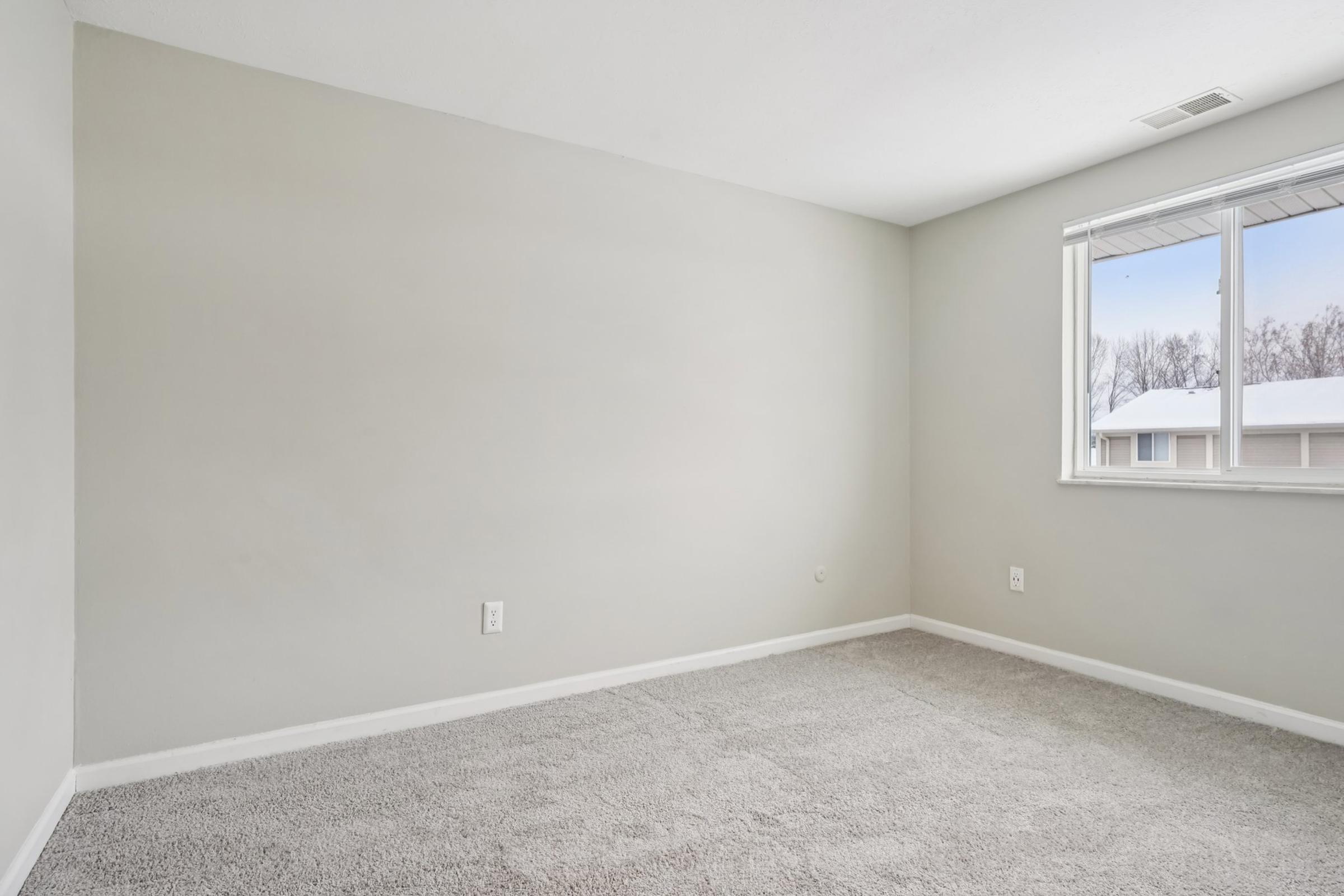 Empty room with light gray walls and a large window letting in natural light. The floor is covered with plush, light gray carpet. There are no furnishings or decorations, creating a minimalist and spacious feel.