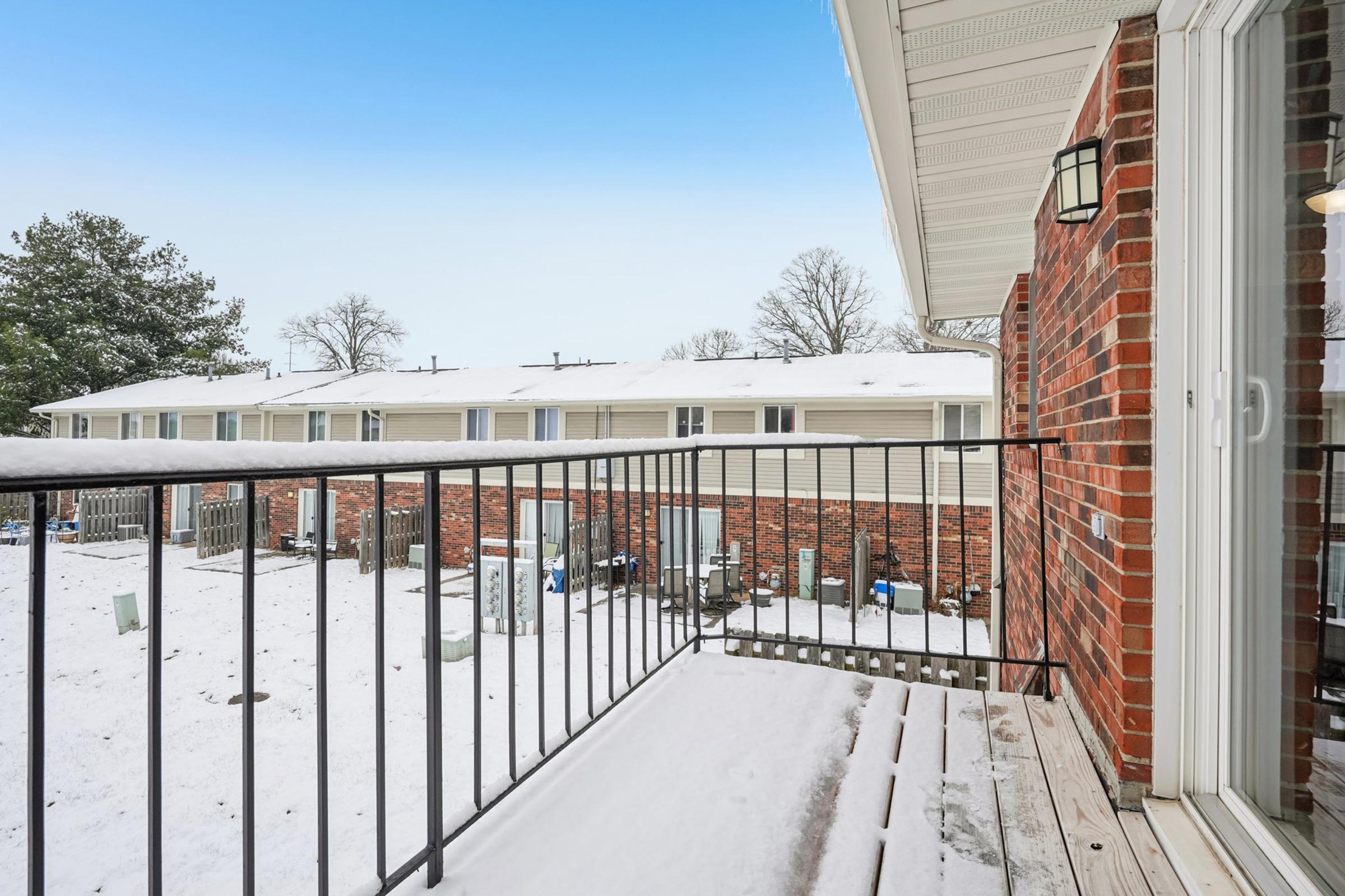 View from a balcony overlooking a snowy courtyard. The scene features several brick apartment buildings in the background, with a clear blue sky above. Snow covers the ground and balcony railing, creating a peaceful winter atmosphere.