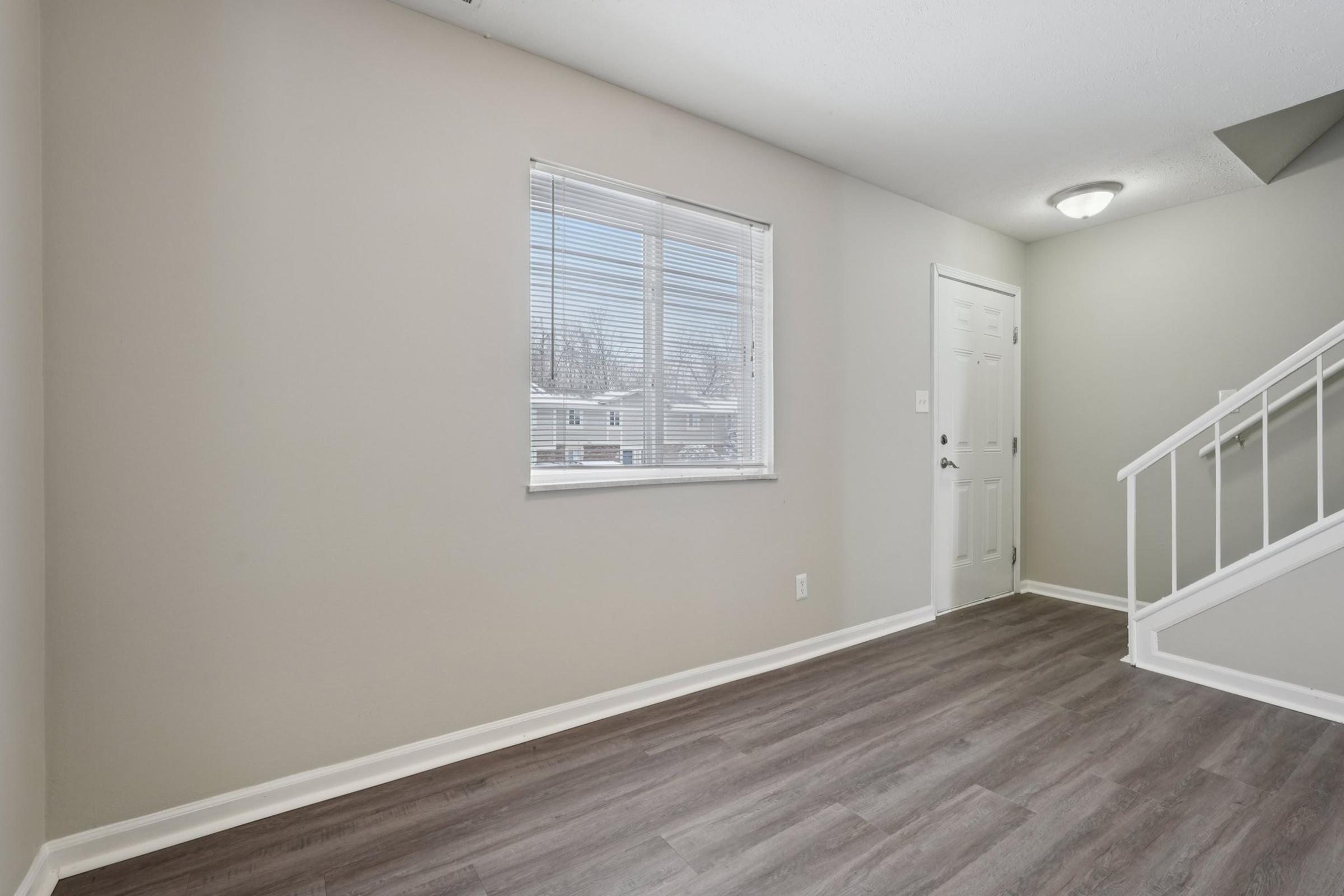 A spacious, light-filled room with gray walls and a large window allowing natural light in. There is a white door leading outside and a staircase on the right, with wooden flooring adding warmth to the space. The overall design is modern and minimalistic.