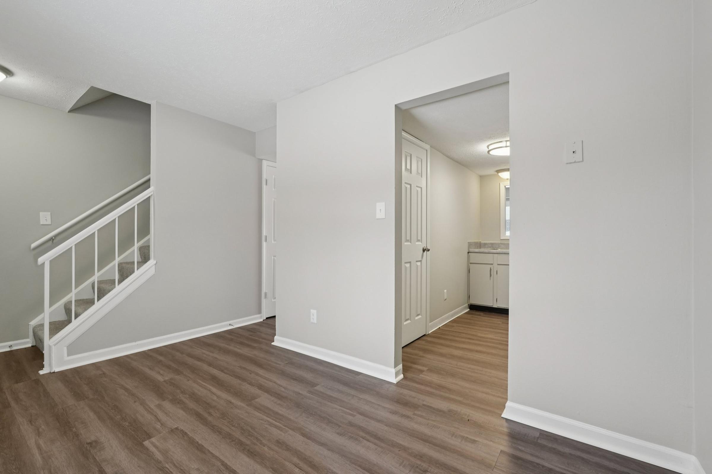 Interior view of a modern, newly renovated home featuring light gray walls and dark wood-like flooring. A staircase is visible on the left, and an open doorway leads to a small bathroom in the background. Natural light comes from a window in the bathroom area, creating a bright and inviting atmosphere.